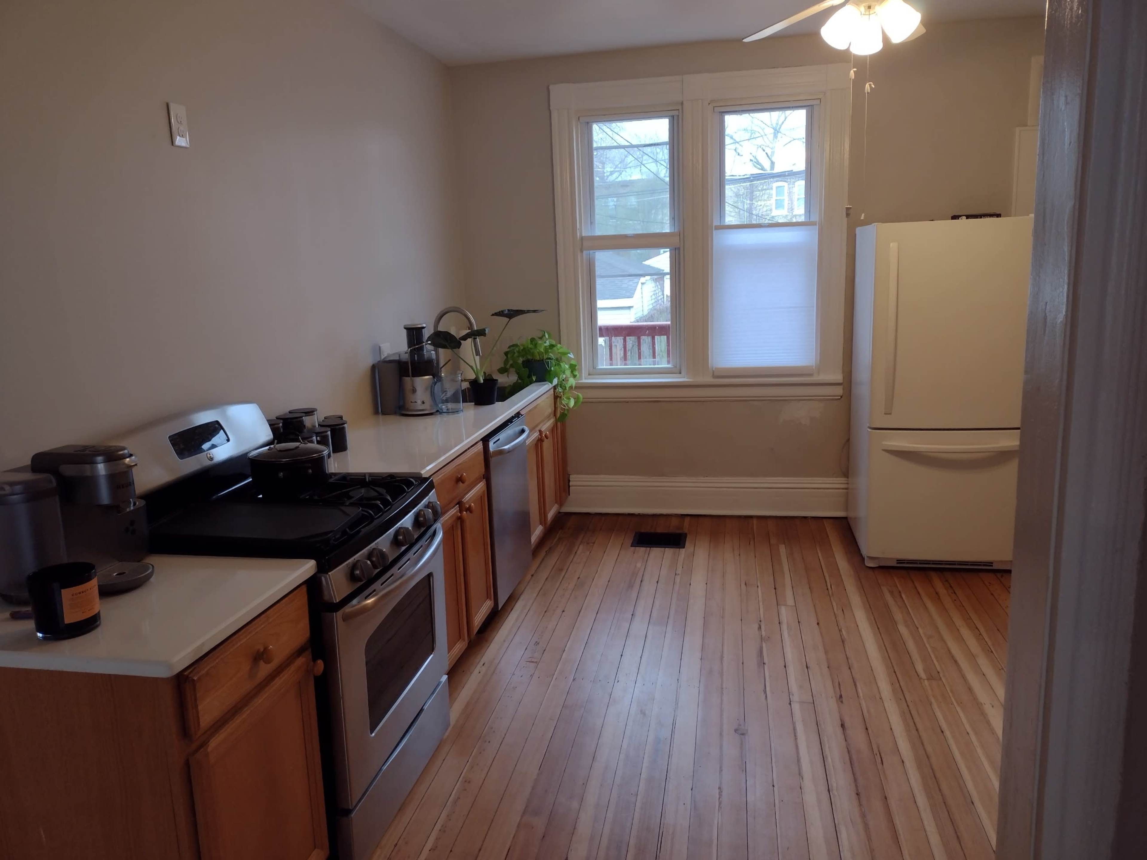 A kitchen with wooden floors, a gas stove, a refrigerator, and a window providing natural light.