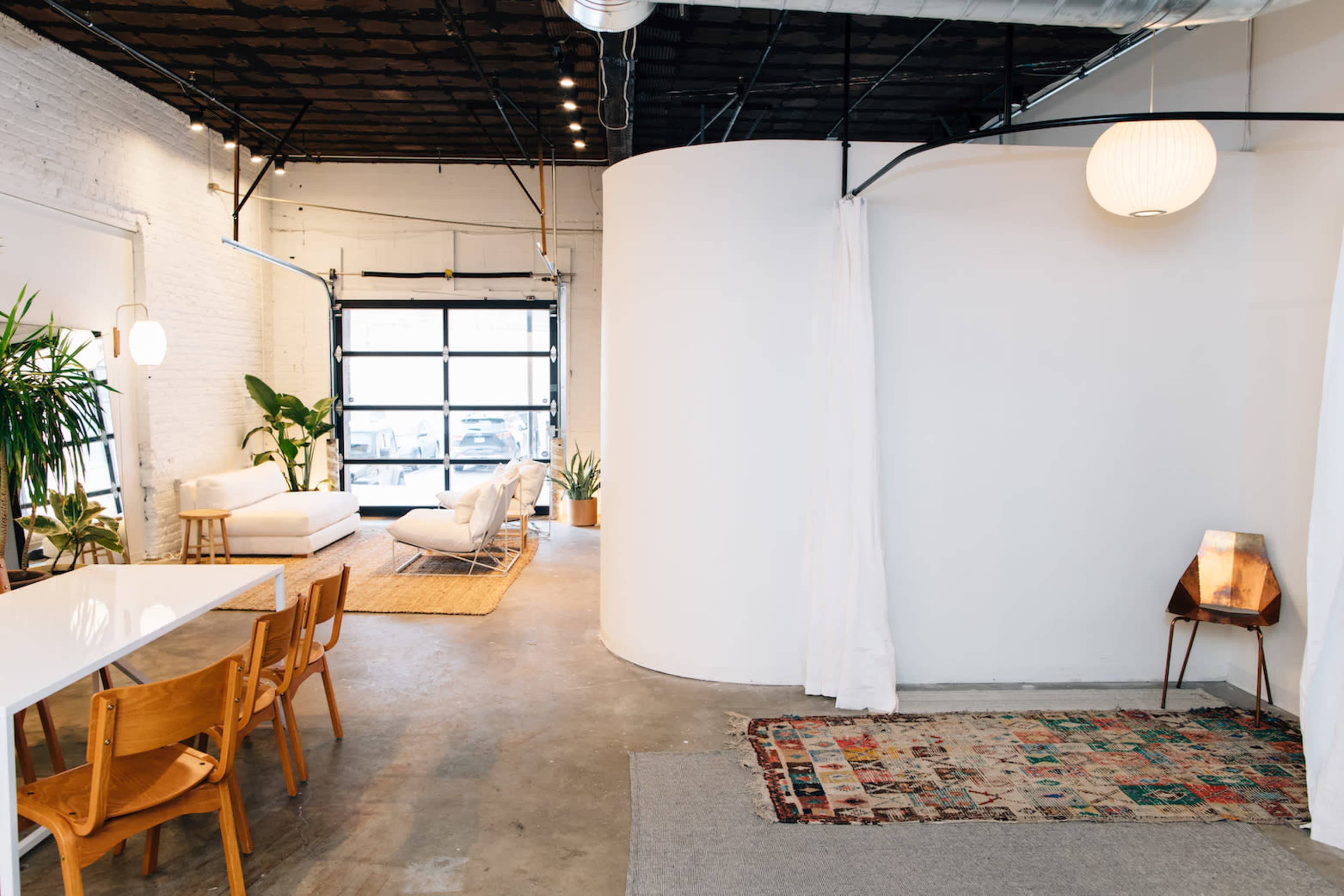 The image shows a spacious interior with a dining area featuring wooden chairs, a round partition, and various plants alongside a cozy seating area illuminated by soft lighting.