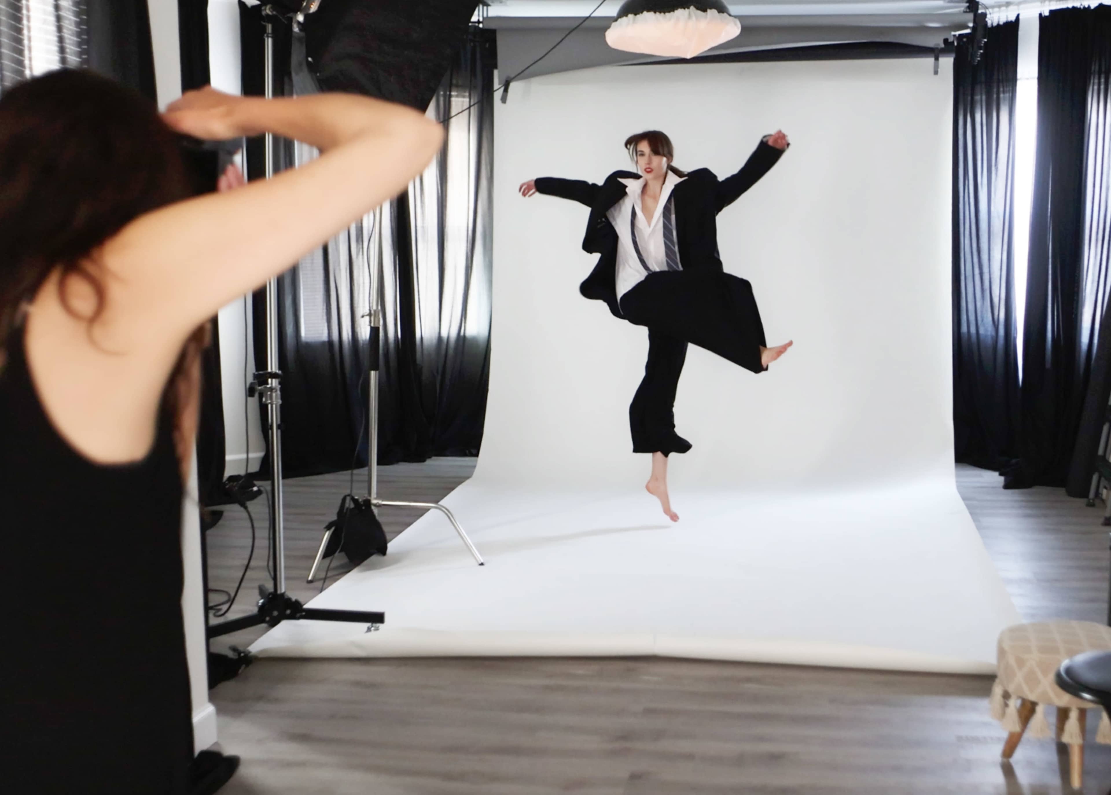 A person in a black suit performs a dance pose on a white backdrop while another individual takes photos.