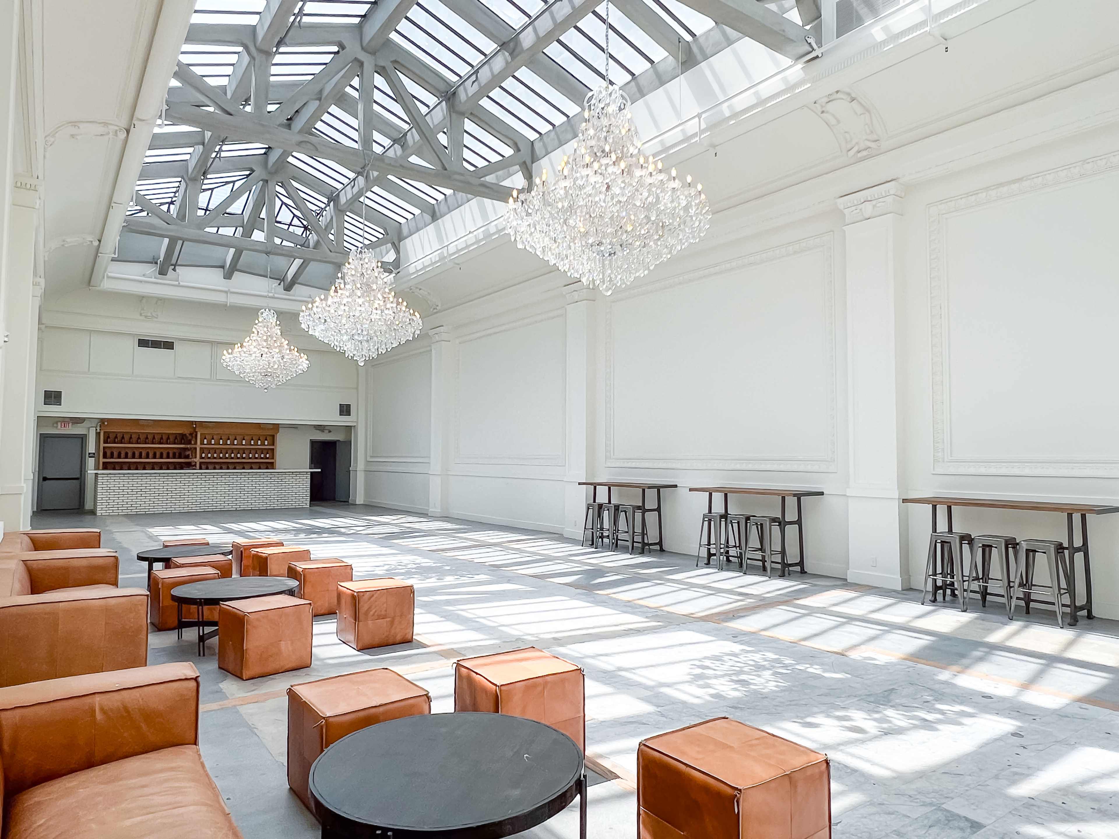 A spacious, well-lit room with large windows, featuring several crystal chandeliers, leather seating, and a bar area in the background.