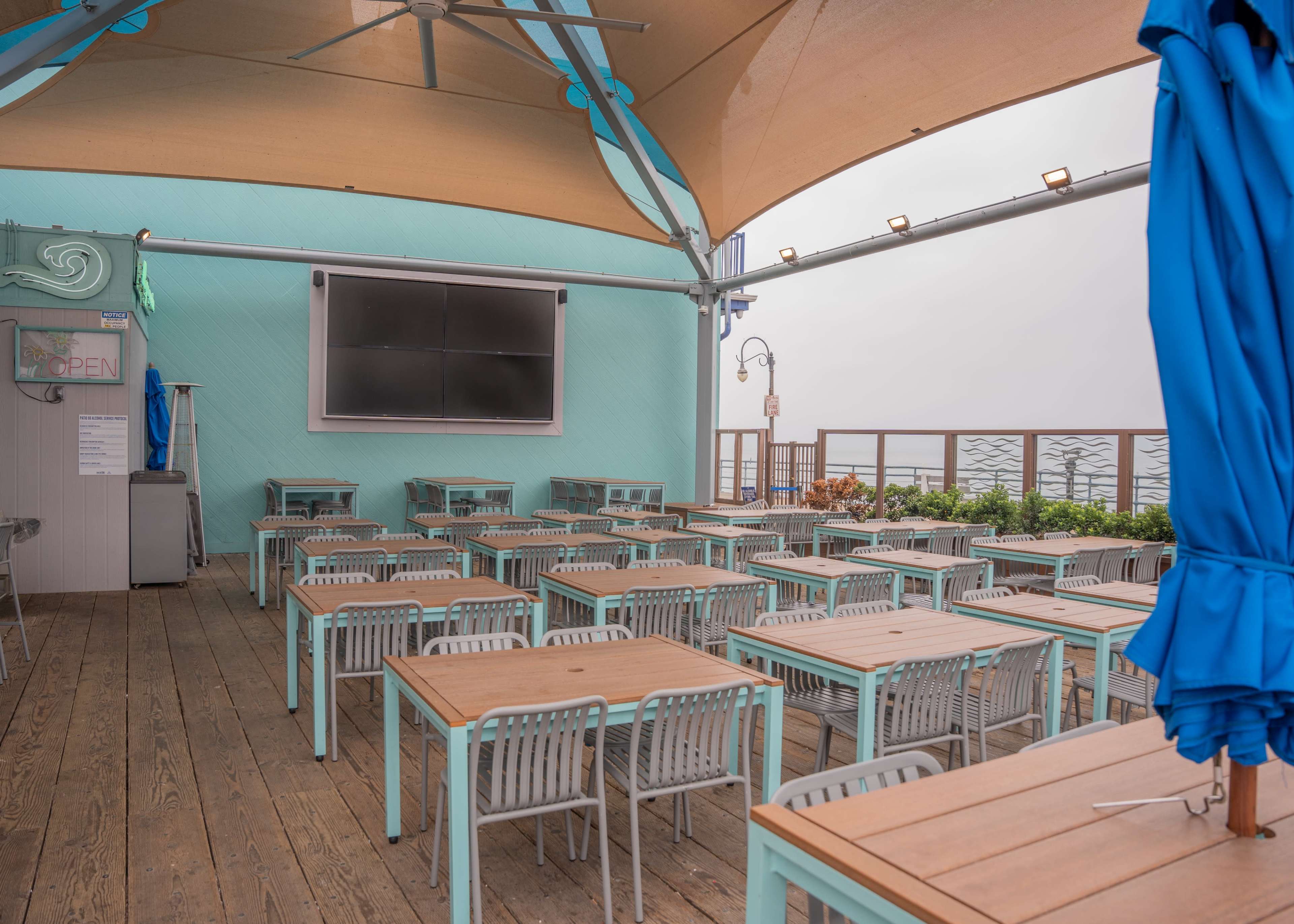The image shows an outdoor dining area with numerous tables and chairs, covered by a large awning, and featuring a mounted television.
