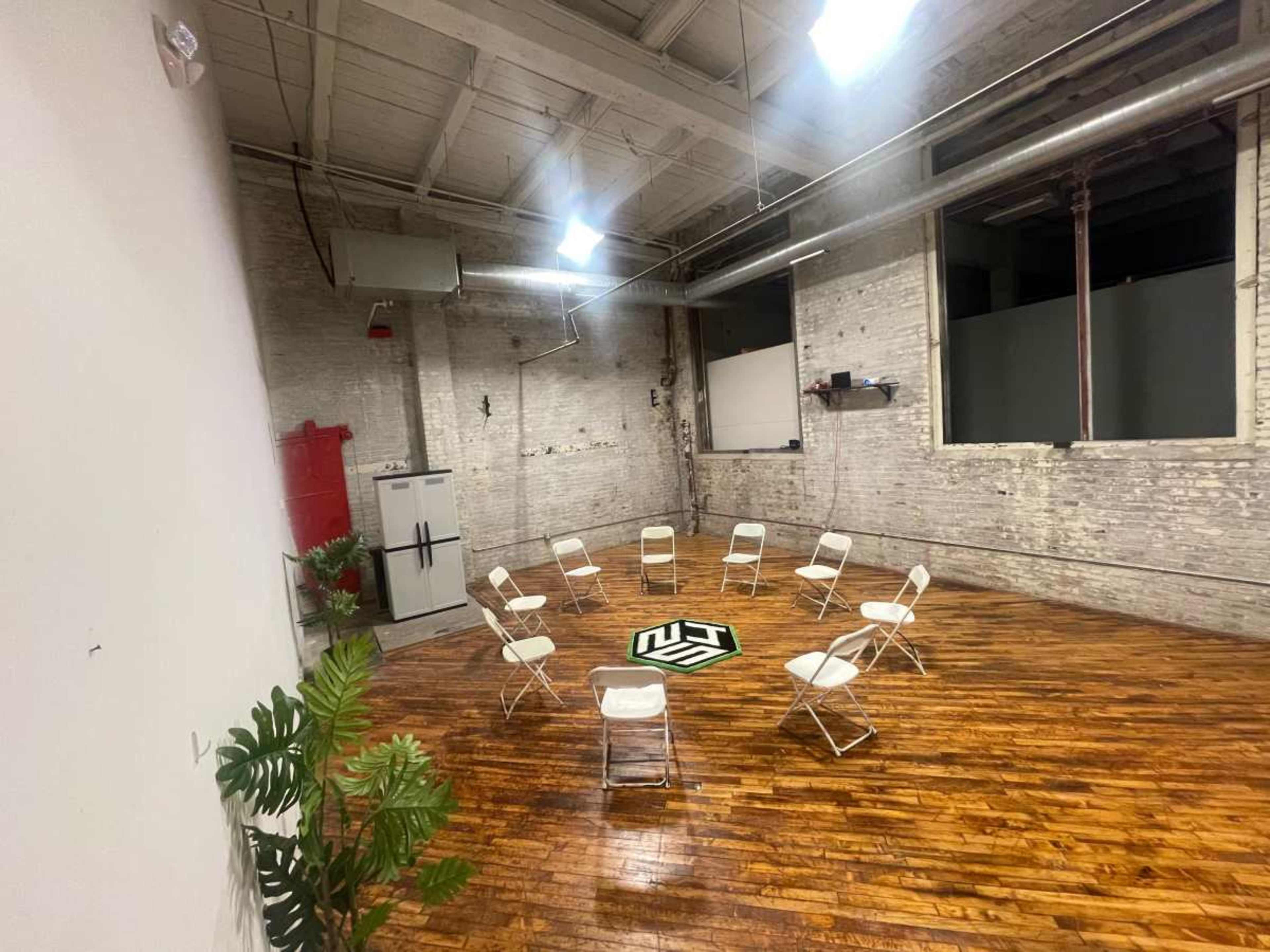 A spacious room with wooden floors features a circle of white folding chairs and a central logo on the ground, with exposed brick walls and minimal furnishings.