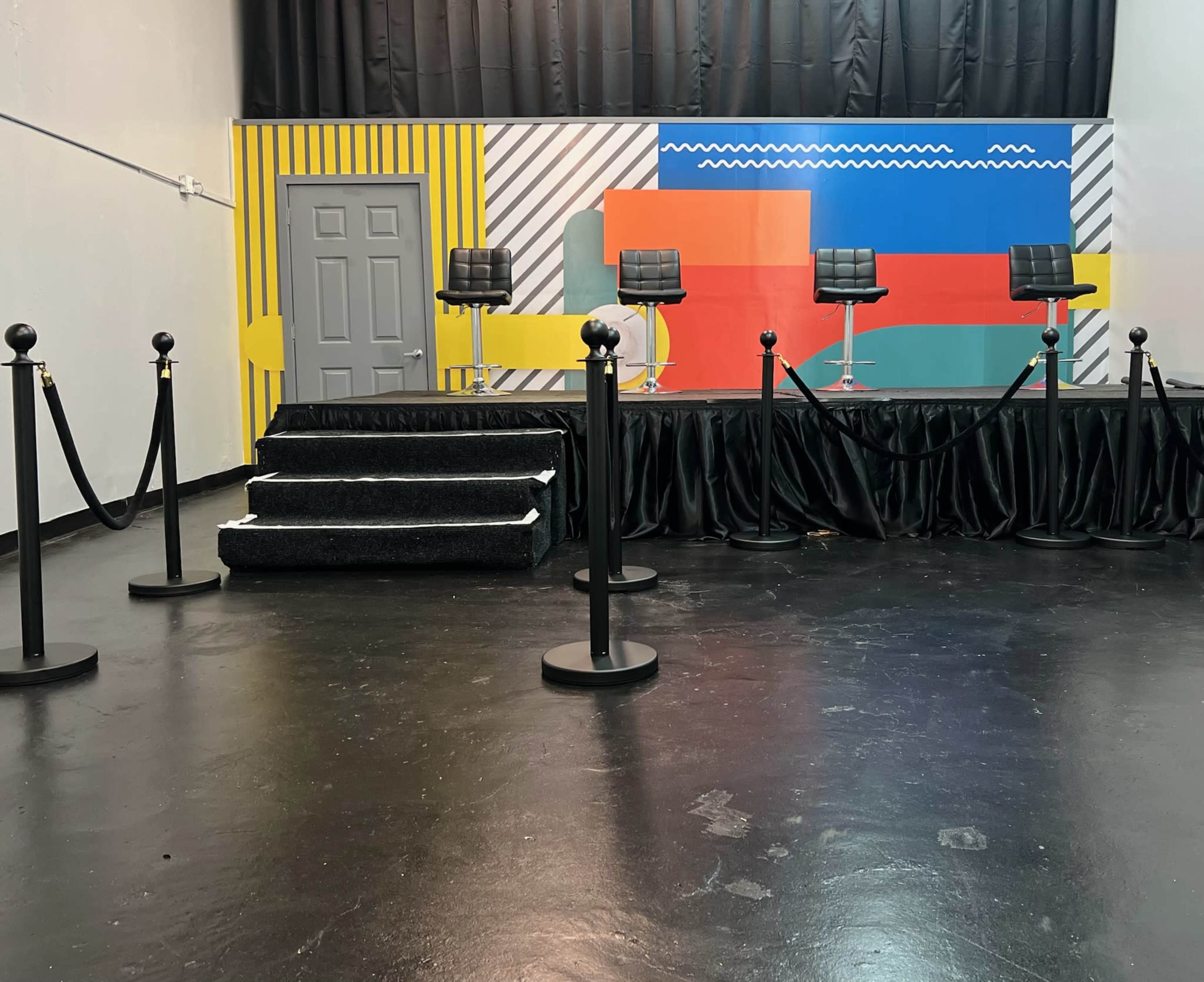 The image shows a staged area with a patterned backdrop, four empty chairs on pedestals, and a set of stanchions with ropes leading to a stairway.