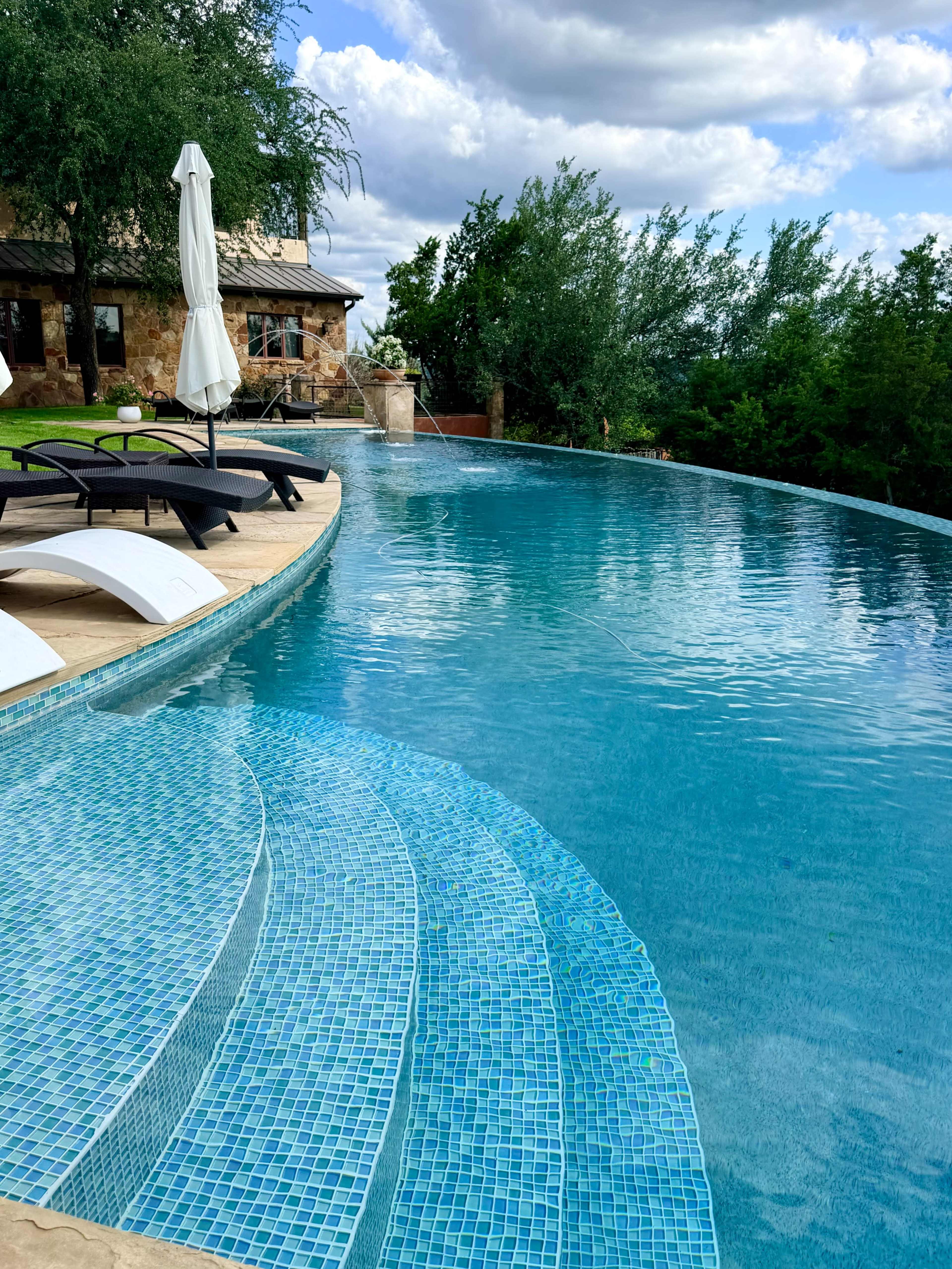 The image shows a blue infinity pool with a tiled entrance, flanked by lounge chairs and surrounded by greenery under a partly cloudy sky.