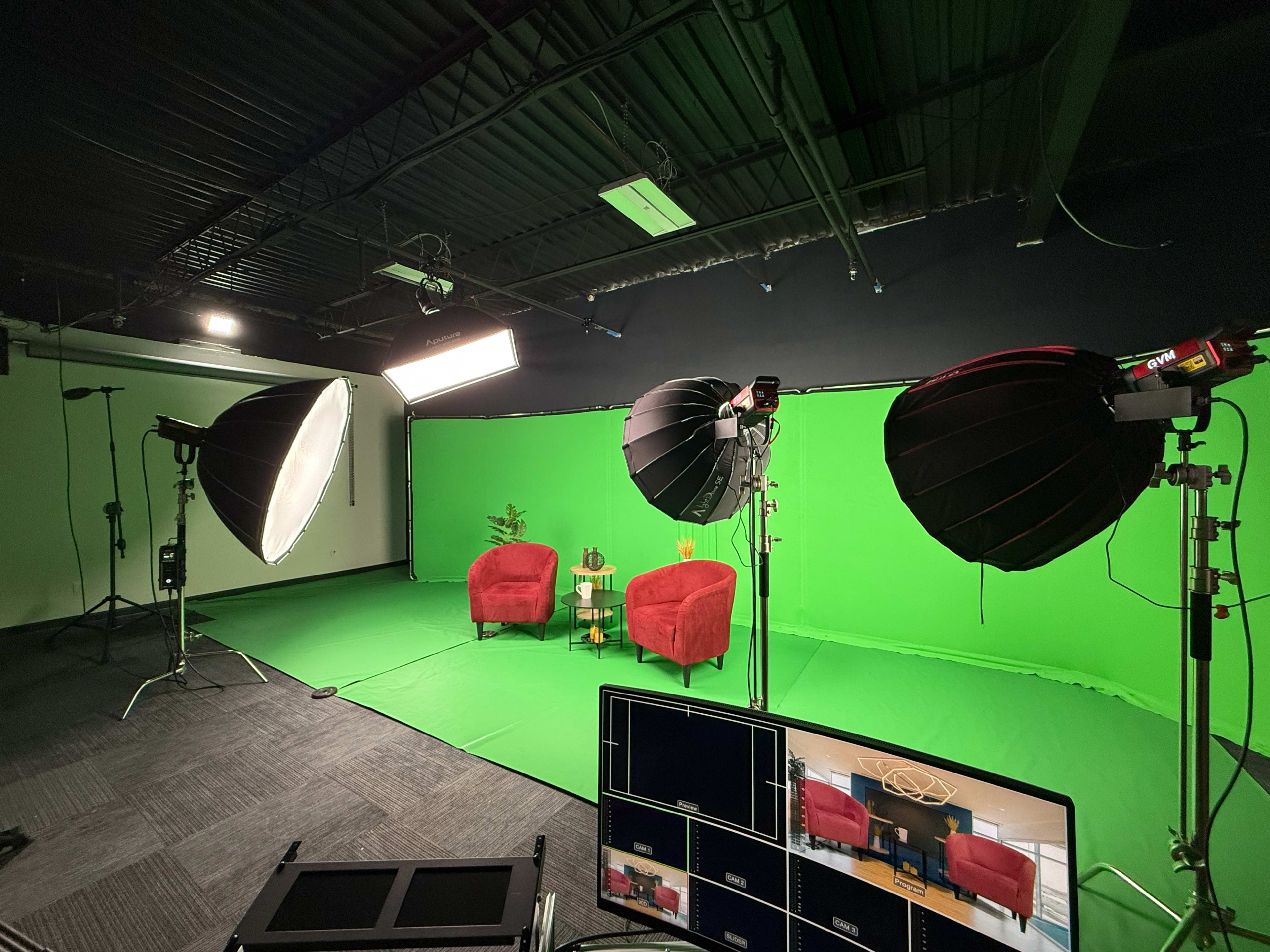 The image shows a television studio setup with two red chairs placed on a green screen, surrounded by professional lighting equipment and a control panel featuring multiple screens.