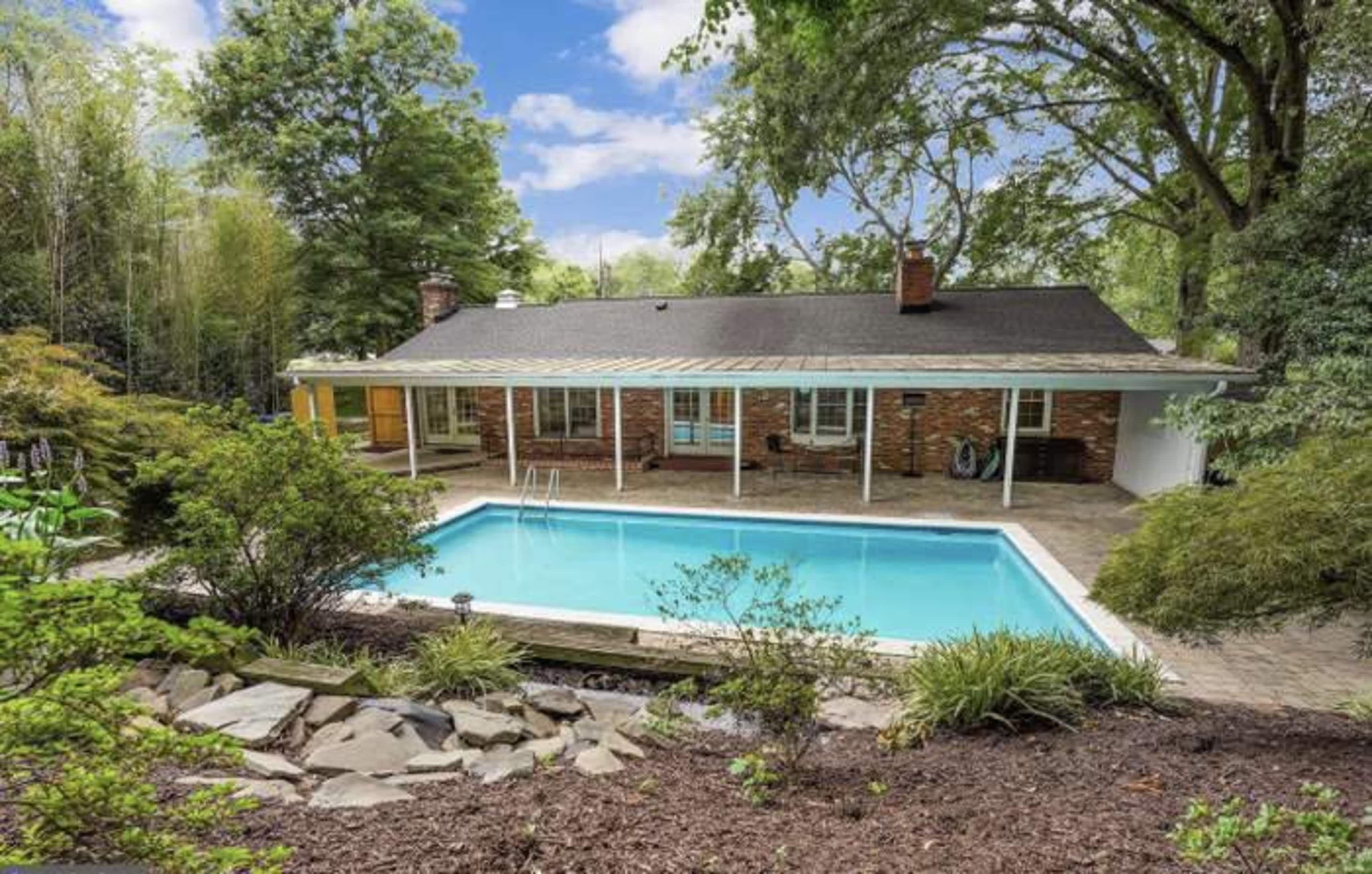 A brick house with a covered patio overlooks a swimming pool surrounded by landscaped gardens and trees.