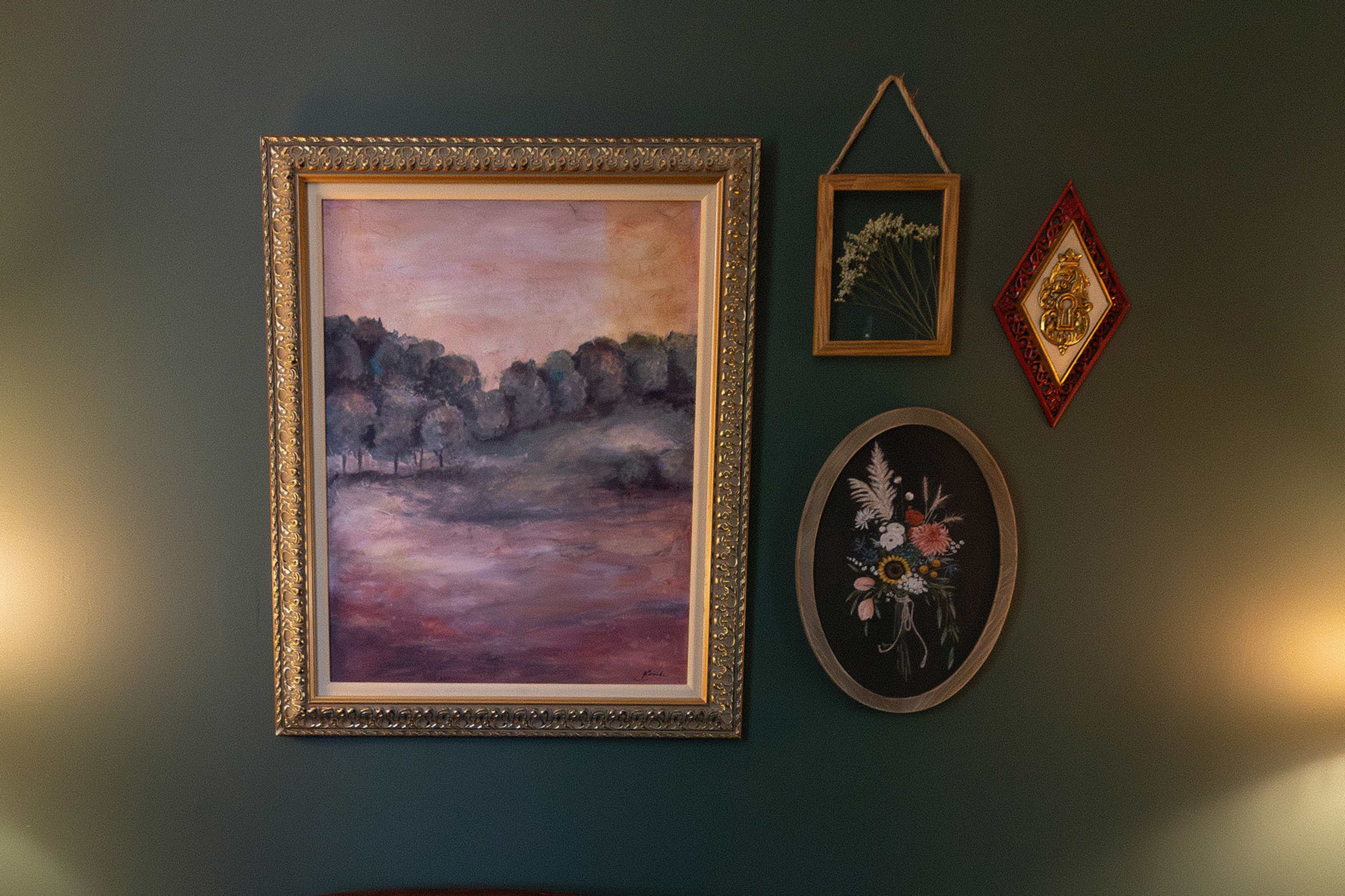 A series of framed artworks arranged on a green wall, including a large landscape painting, a small nature print, and a floral composition in an oval frame.