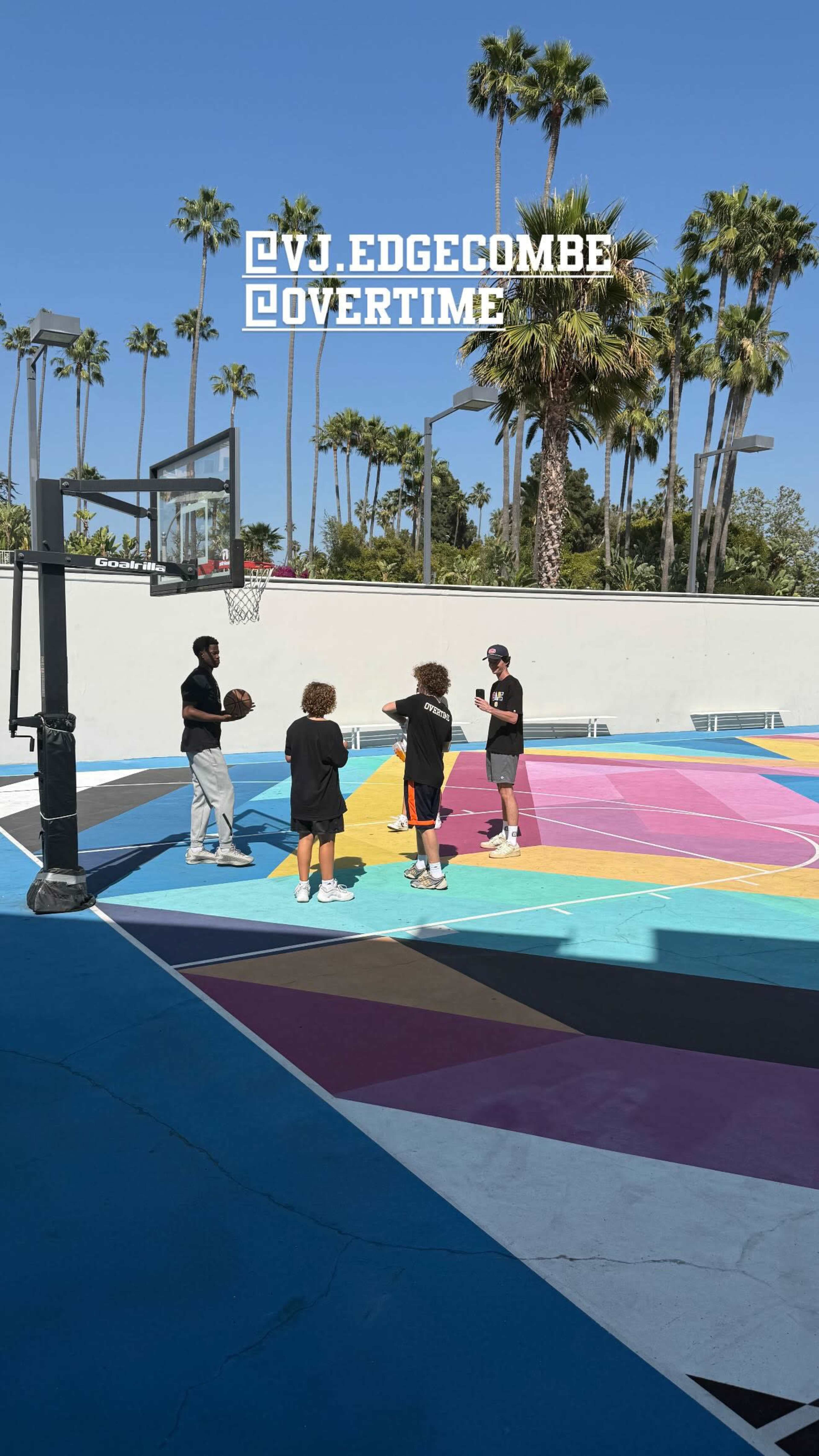 A group of four individuals is gathered on a brightly colored basketball court, speaking with a coach near a basketball hoop under a clear blue sky.