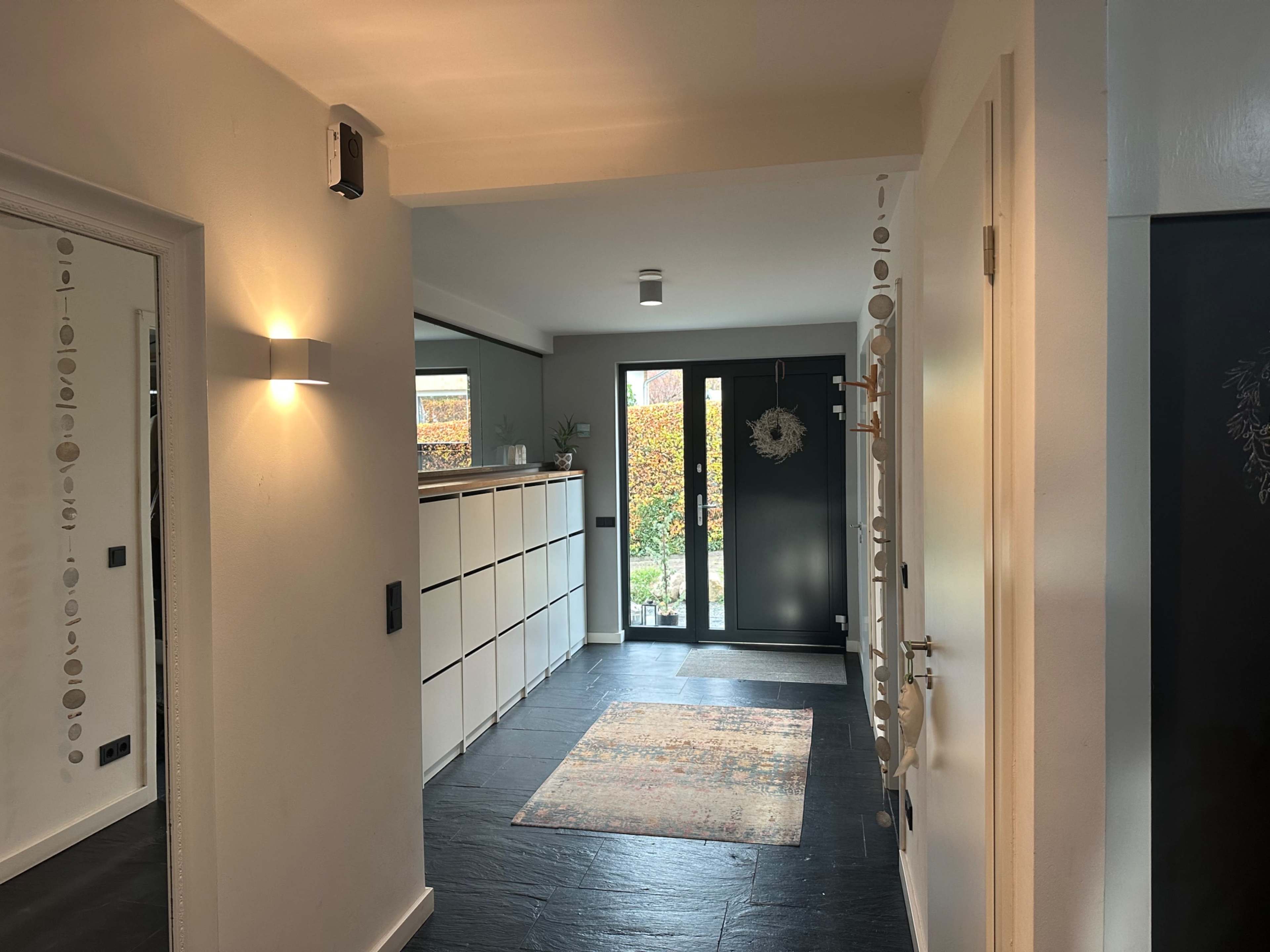 The image shows a well-lit hallway featuring a contemporary design with a rug, a console table, and large windows overlooking a garden.