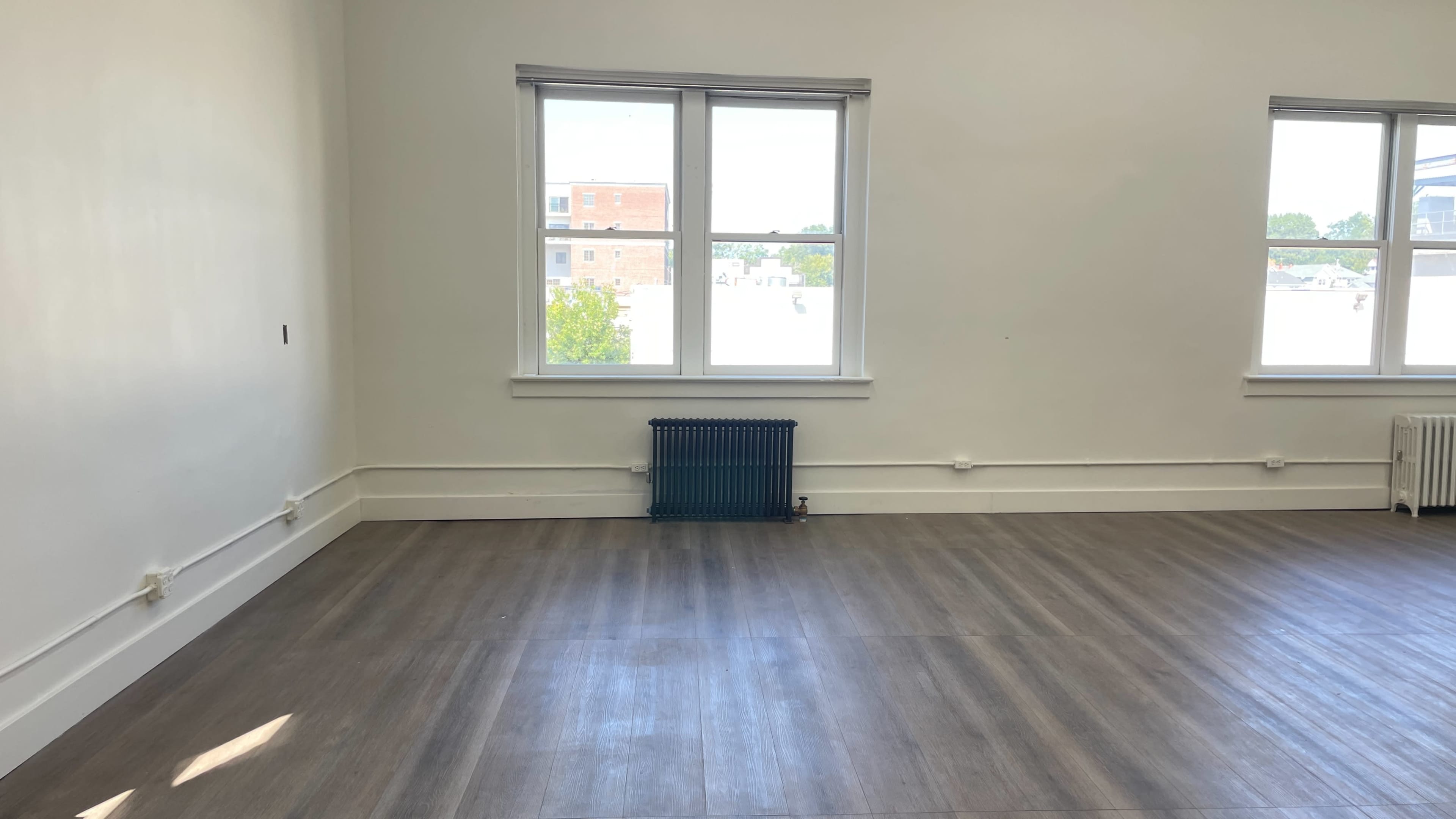 An empty room with light-colored walls, wooden flooring, two windows, and a blue radiator against one wall.
