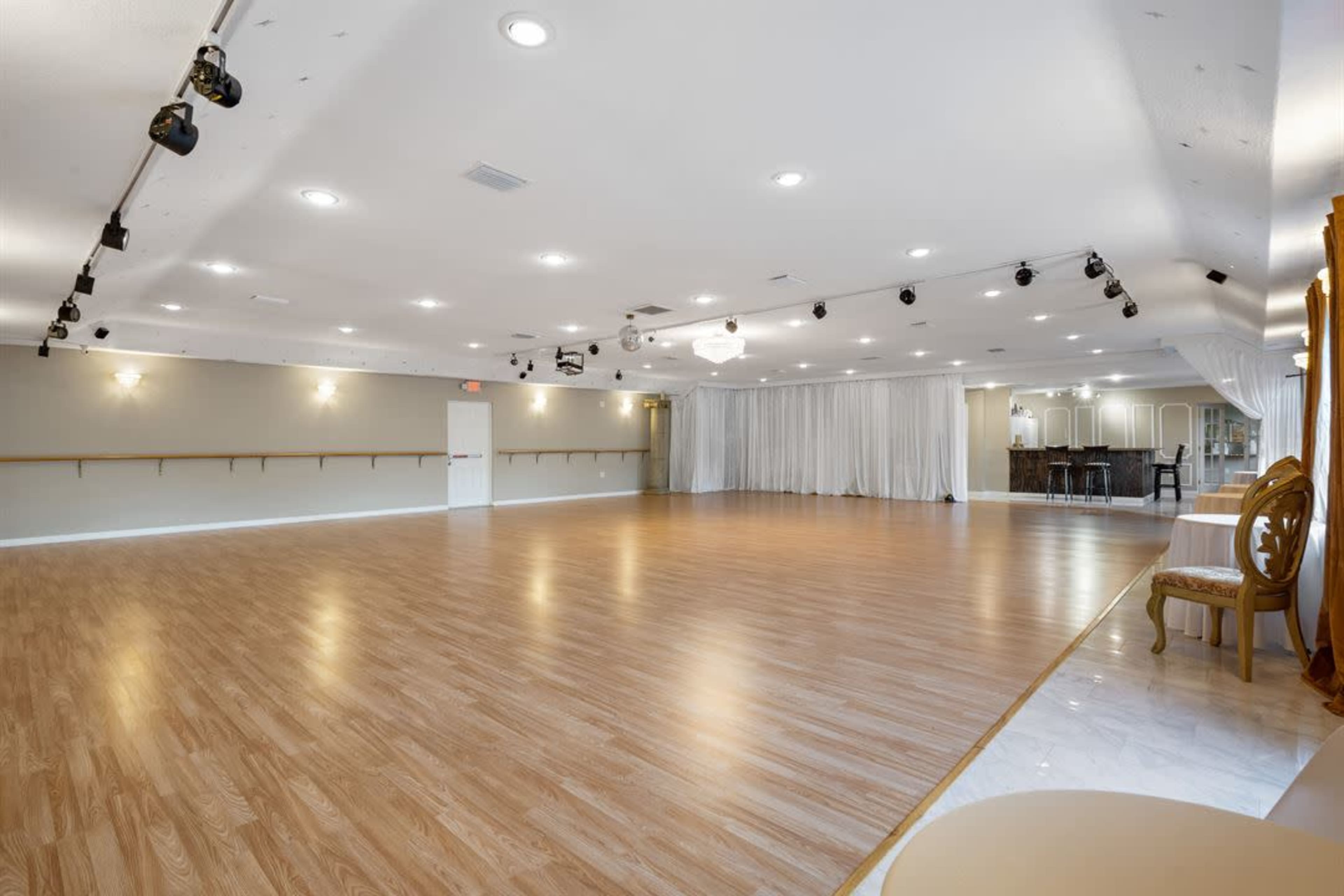 A spacious, well-lit dance studio with polished wooden floors and a mirrored wall, equipped with overhead lighting and a small refreshment area.