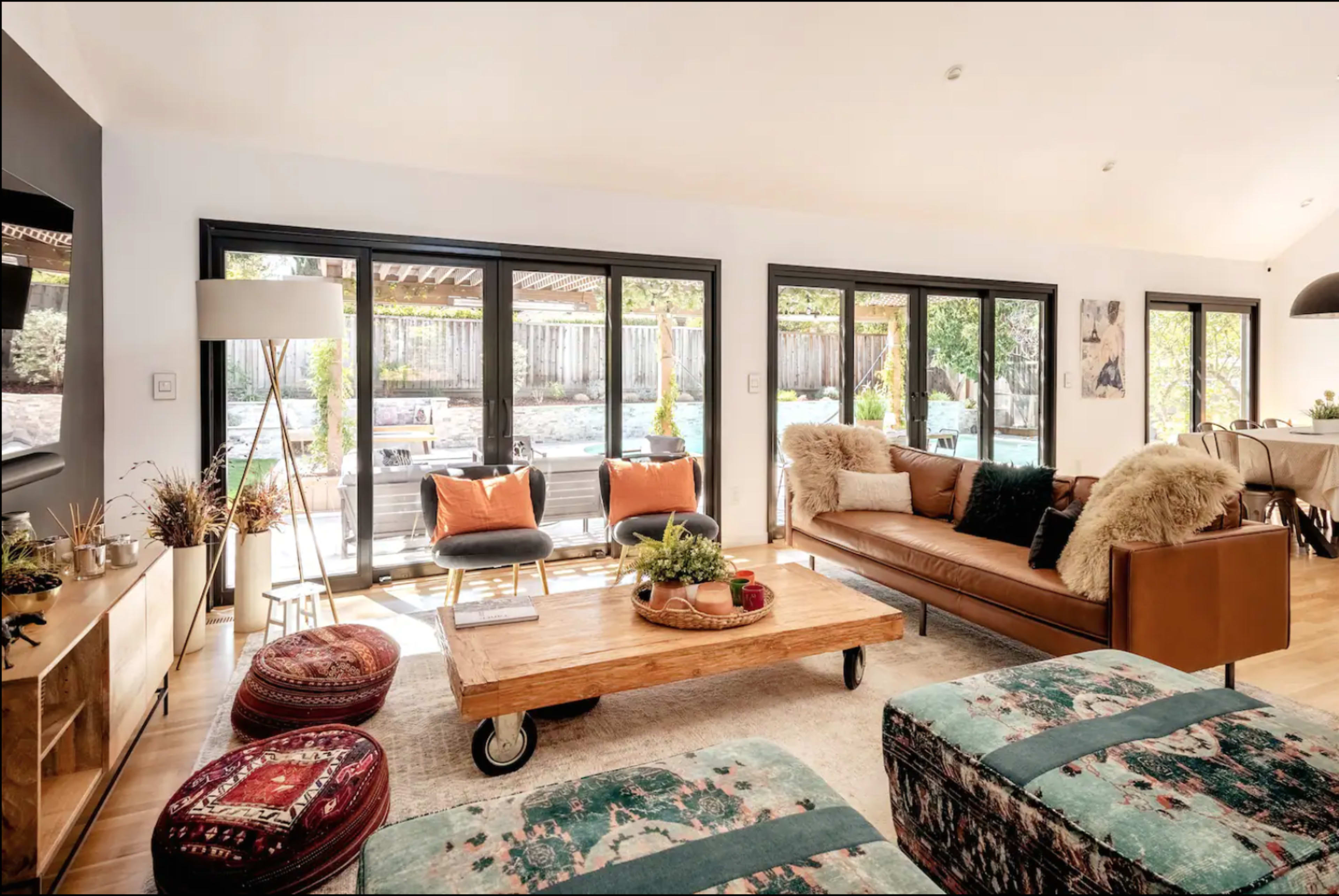A modern living room features a brown leather sofa, an oversized wooden coffee table on wheels, and large glass doors that open to a backyard with a pool.