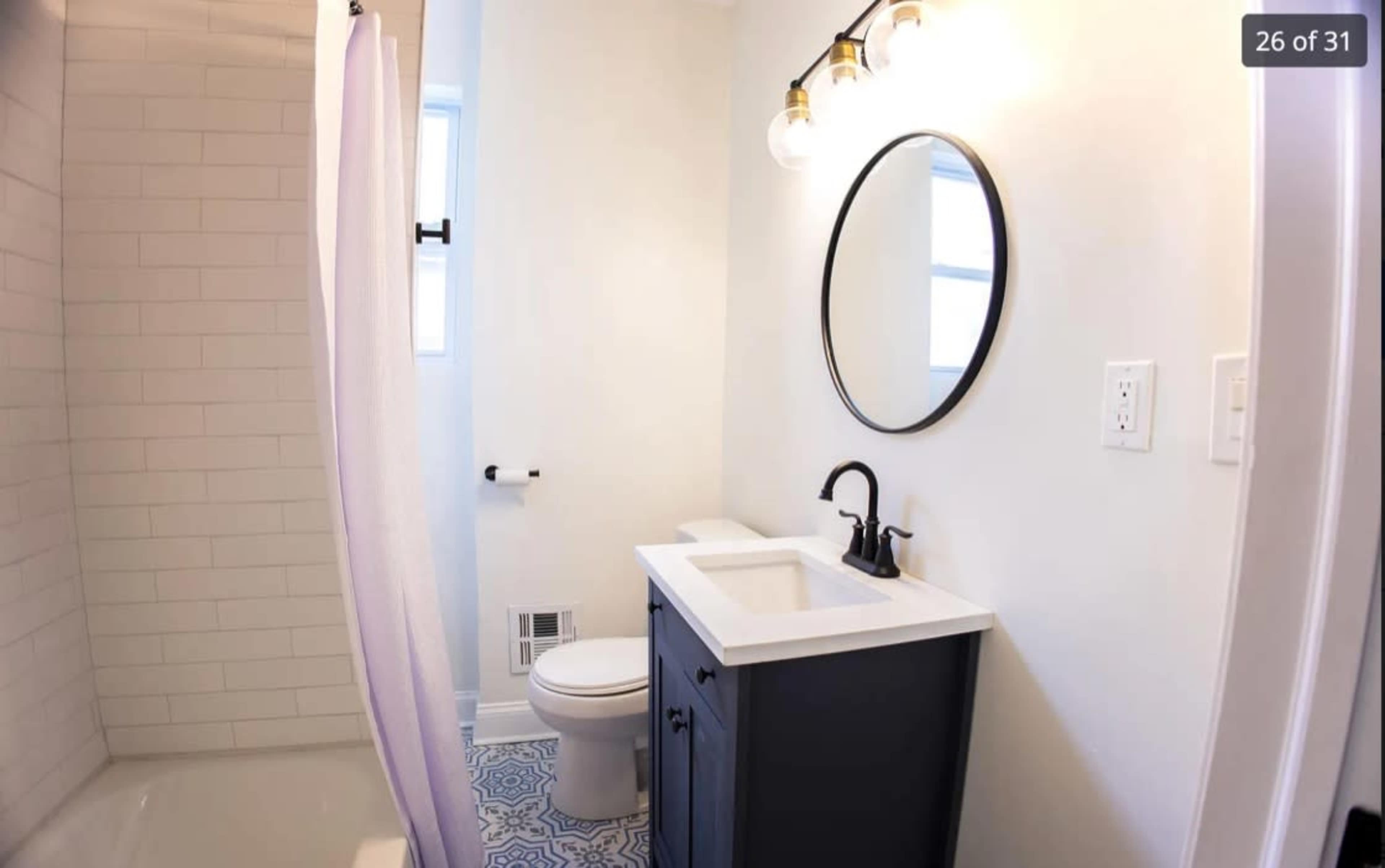 A small bathroom featuring a shower, toilet, and a sink with a dark cabinet, illuminated by three light bulbs and a round mirror above the sink.