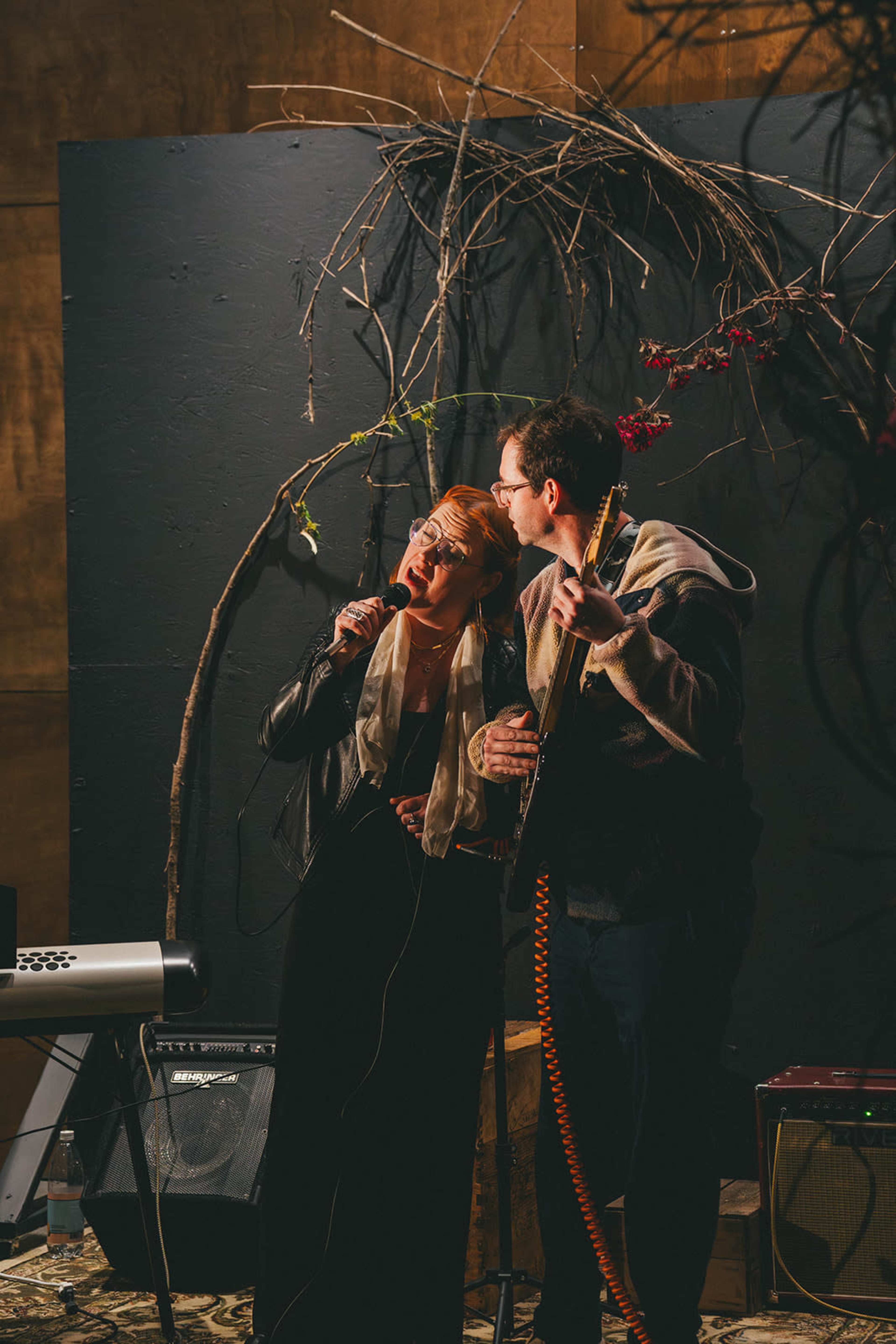 A woman in a black dress and a man in a hoodie sing together on a small stage decorated with branches and flowers.