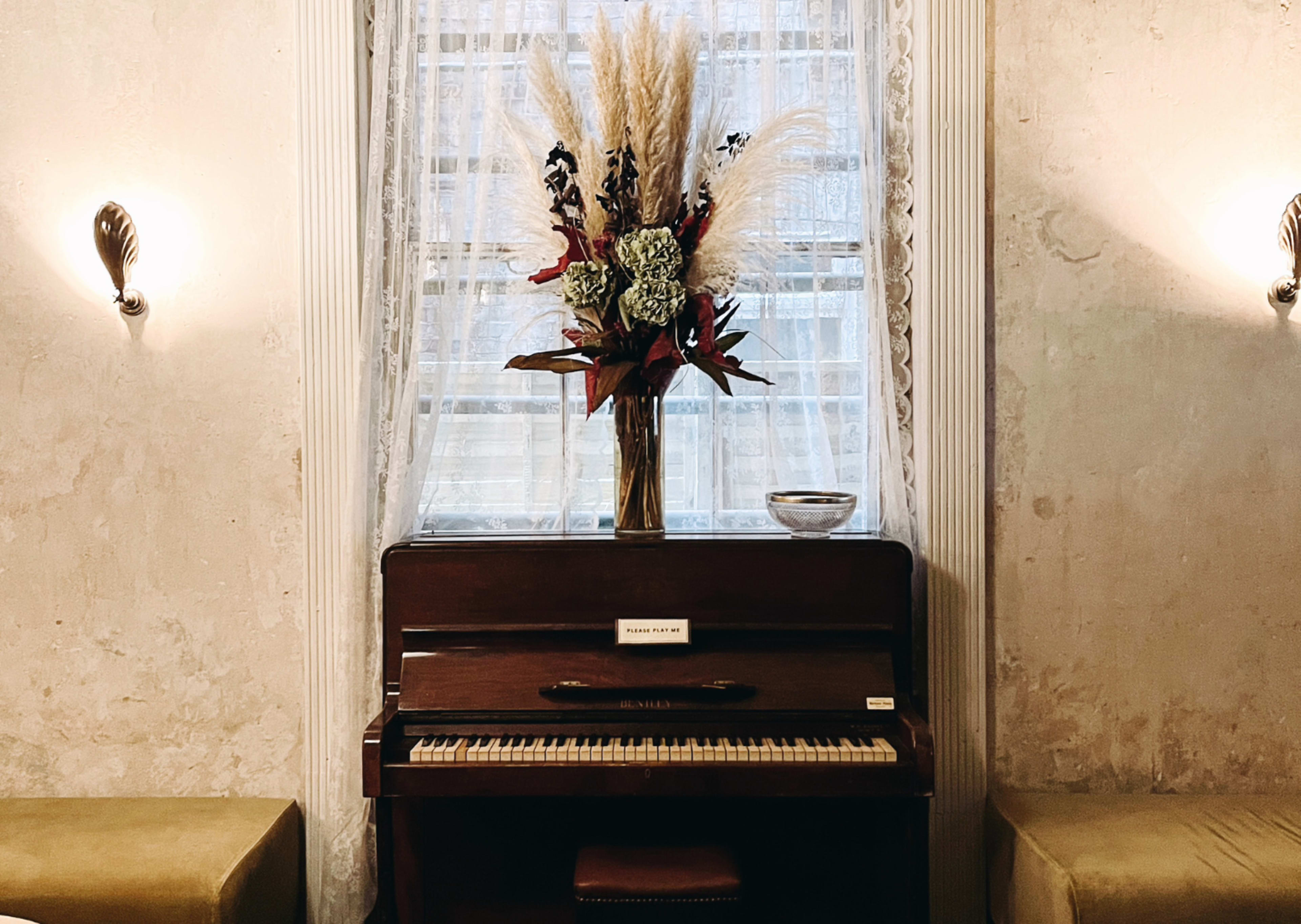 A wooden piano is positioned against a wall, decorated with a vase of dried flowers, with soft lighting illuminating the space.