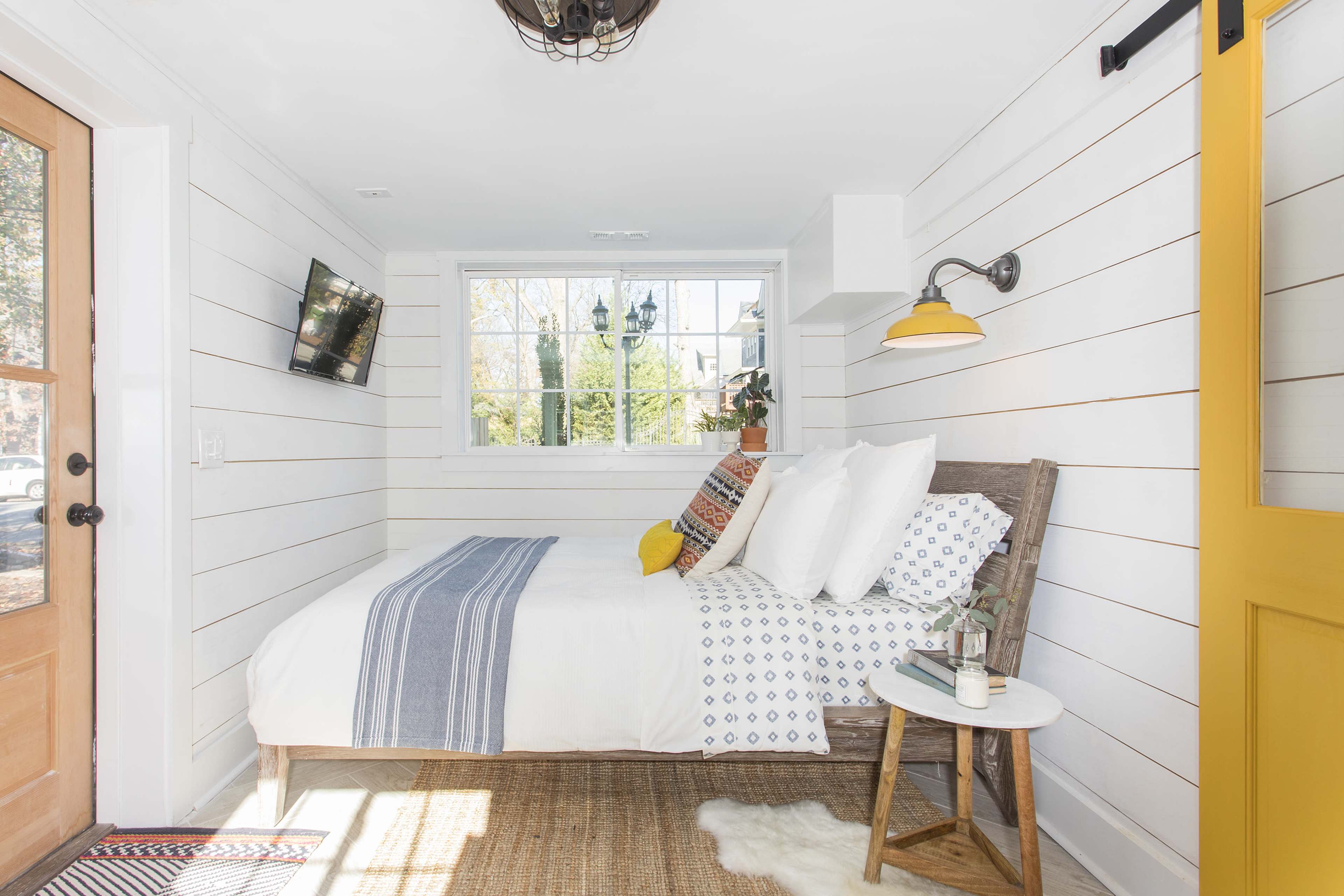 A cozy bedroom with a bed against a wall adorned with white shiplap, featuring a yellow door, a small side table, and a television mounted on the wall.