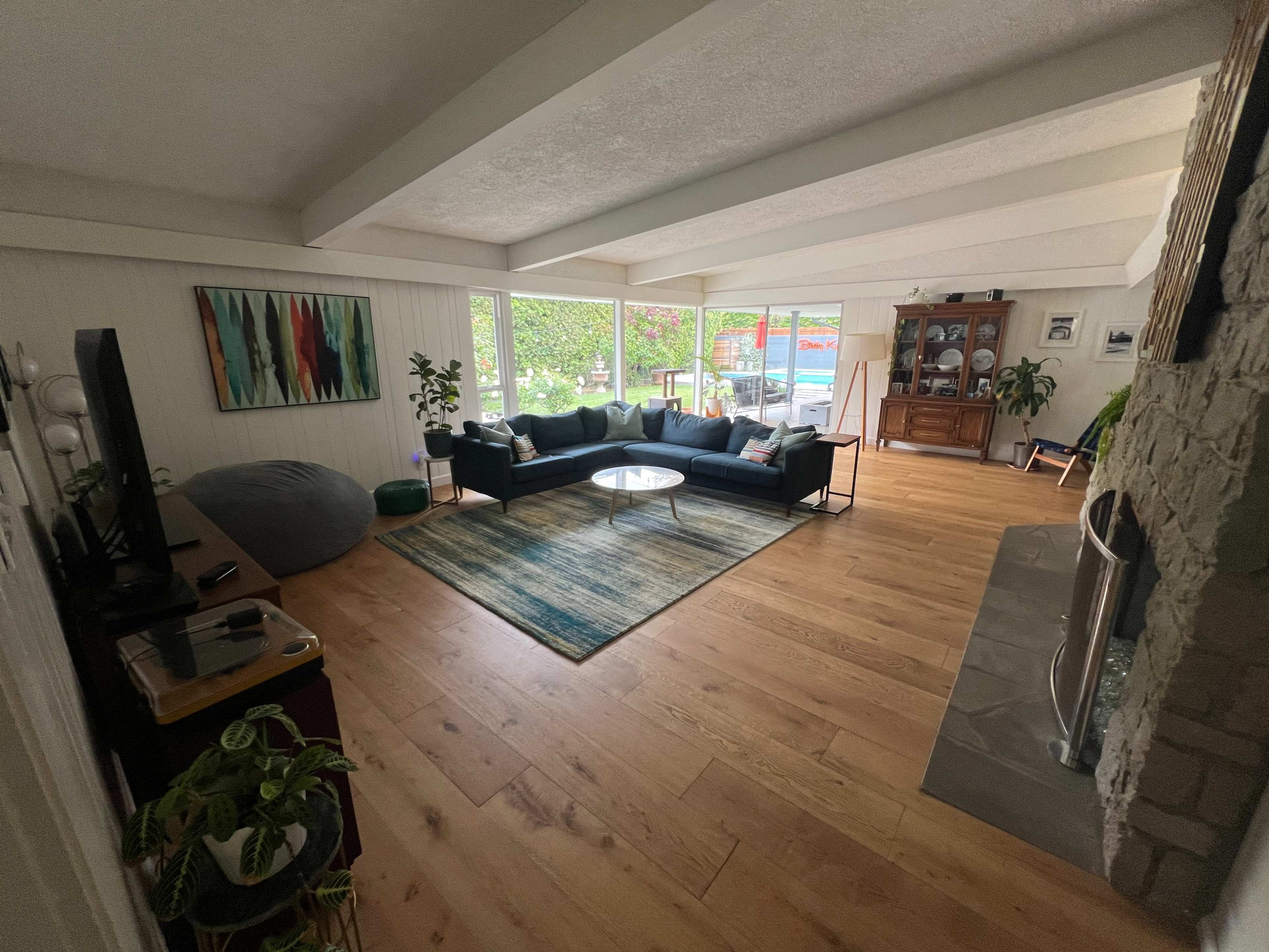 The image shows a spacious living room with a green sofa, a round coffee table, hardwood flooring, and large windows revealing a garden outside.