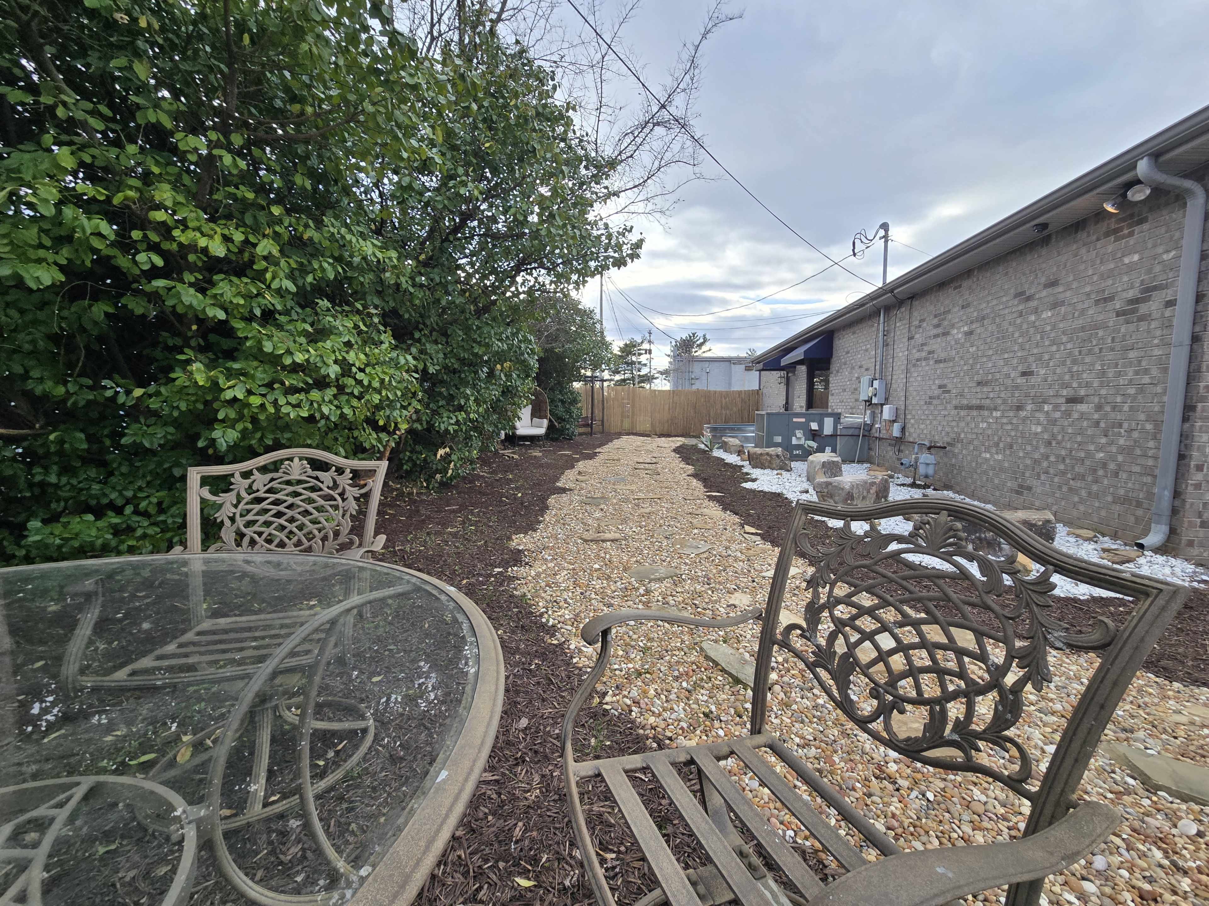 A gravel path lined with mulch and greenery runs alongside a brick building, with a glass-top table and decorative chairs in the foreground.