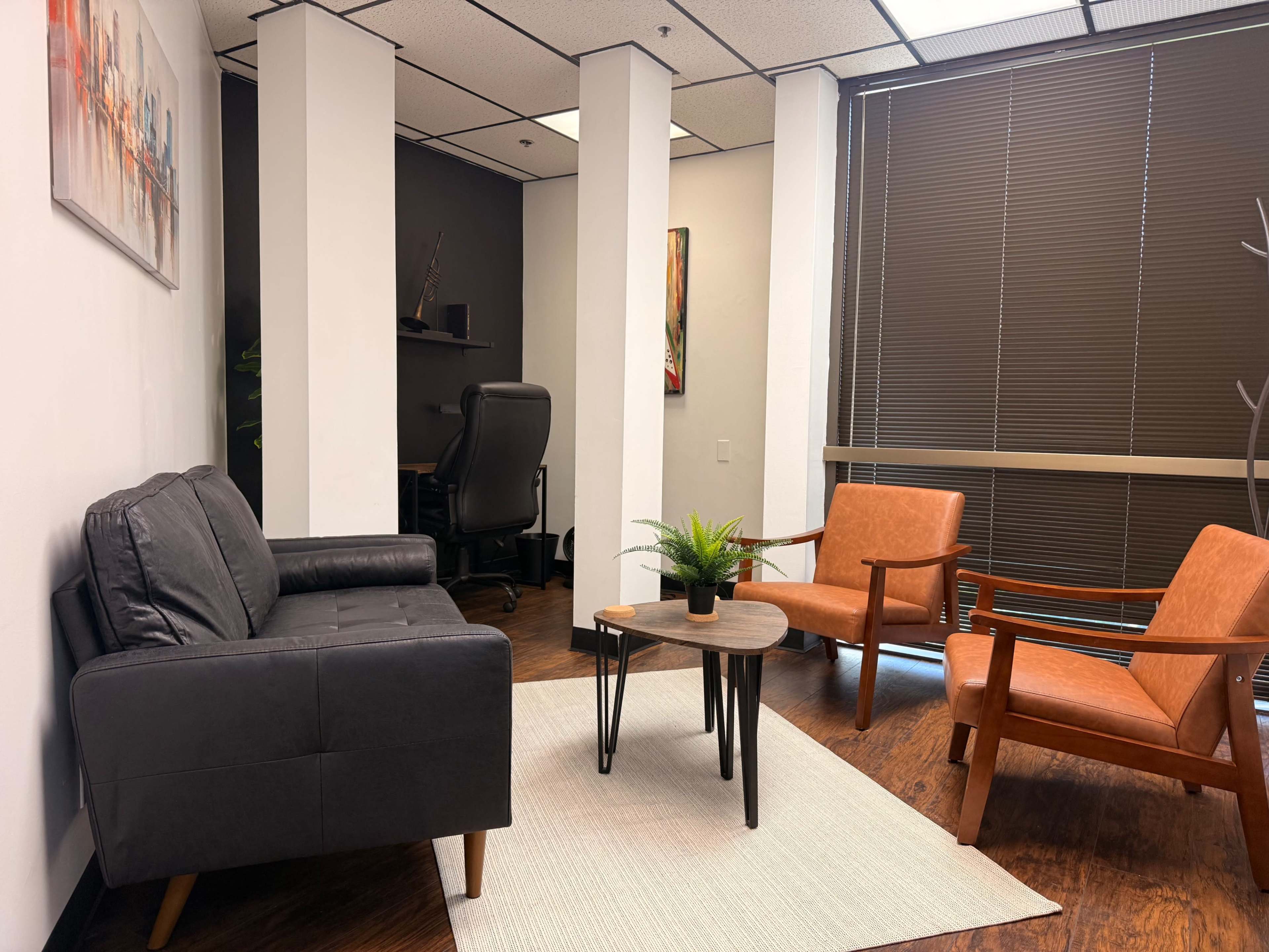 The image shows a modern waiting area with a black couch, two orange armchairs, a small round table, and a desk in the background.