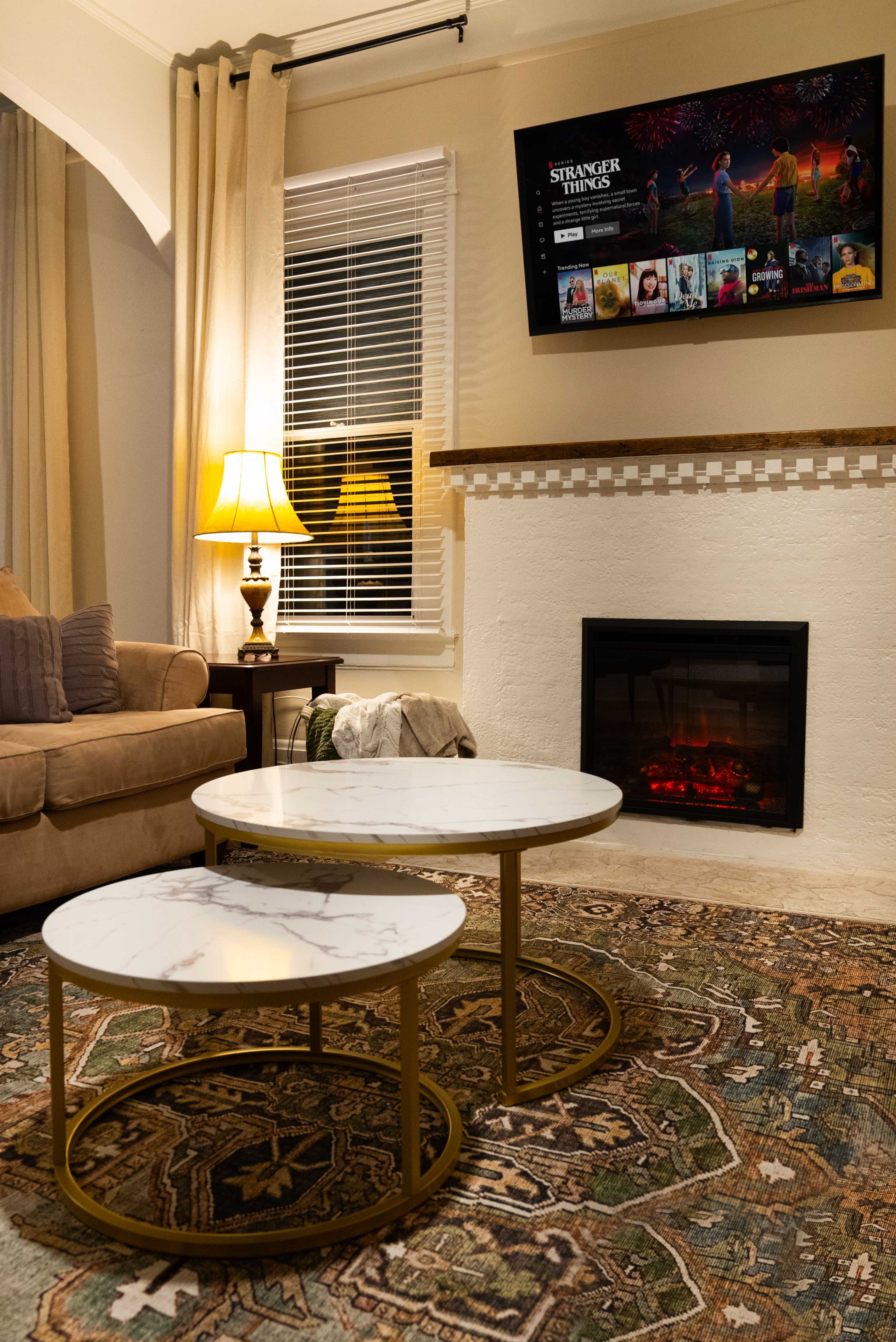 The image shows a cozy living room with a beige sofa, two round marble-top coffee tables, a wall-mounted television displaying "Stranger Things," and a fireplace with an electric flame effect.