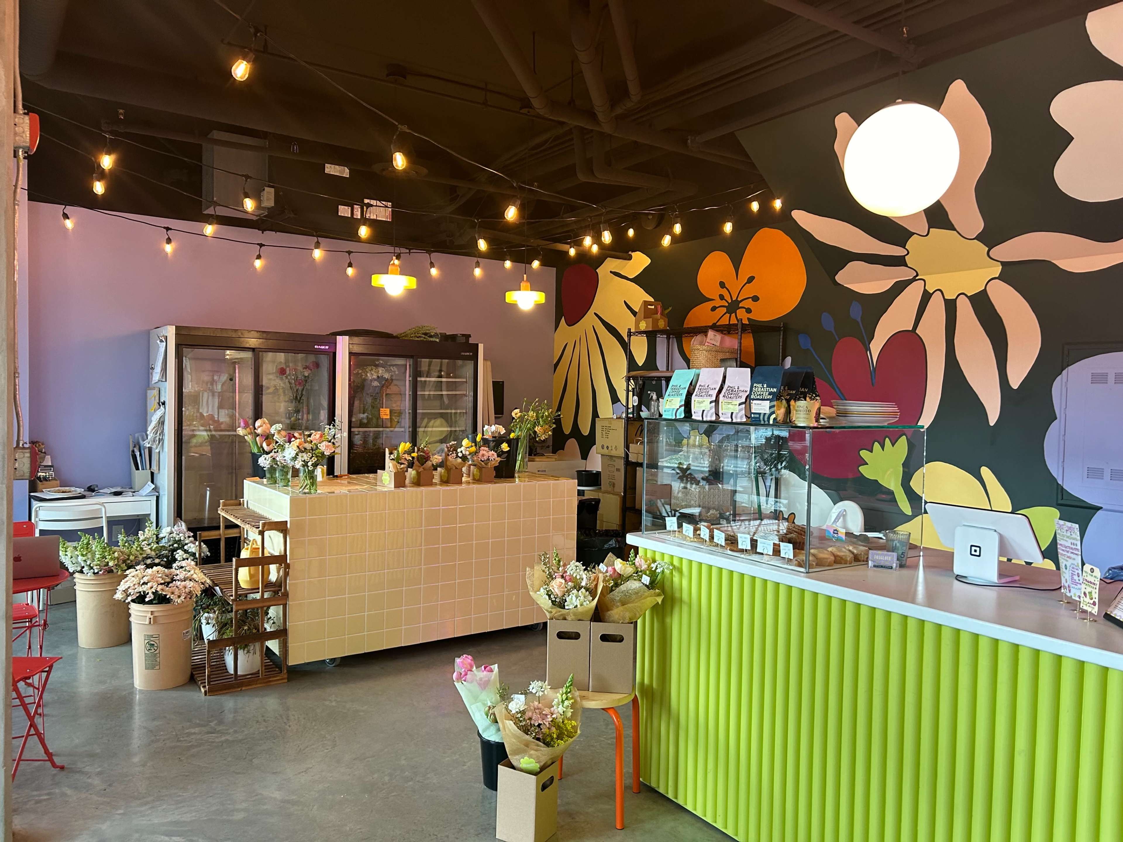 A brightly decorated flower shop interior featuring a colorful mural, display refrigerators, and various floral arrangements on display.