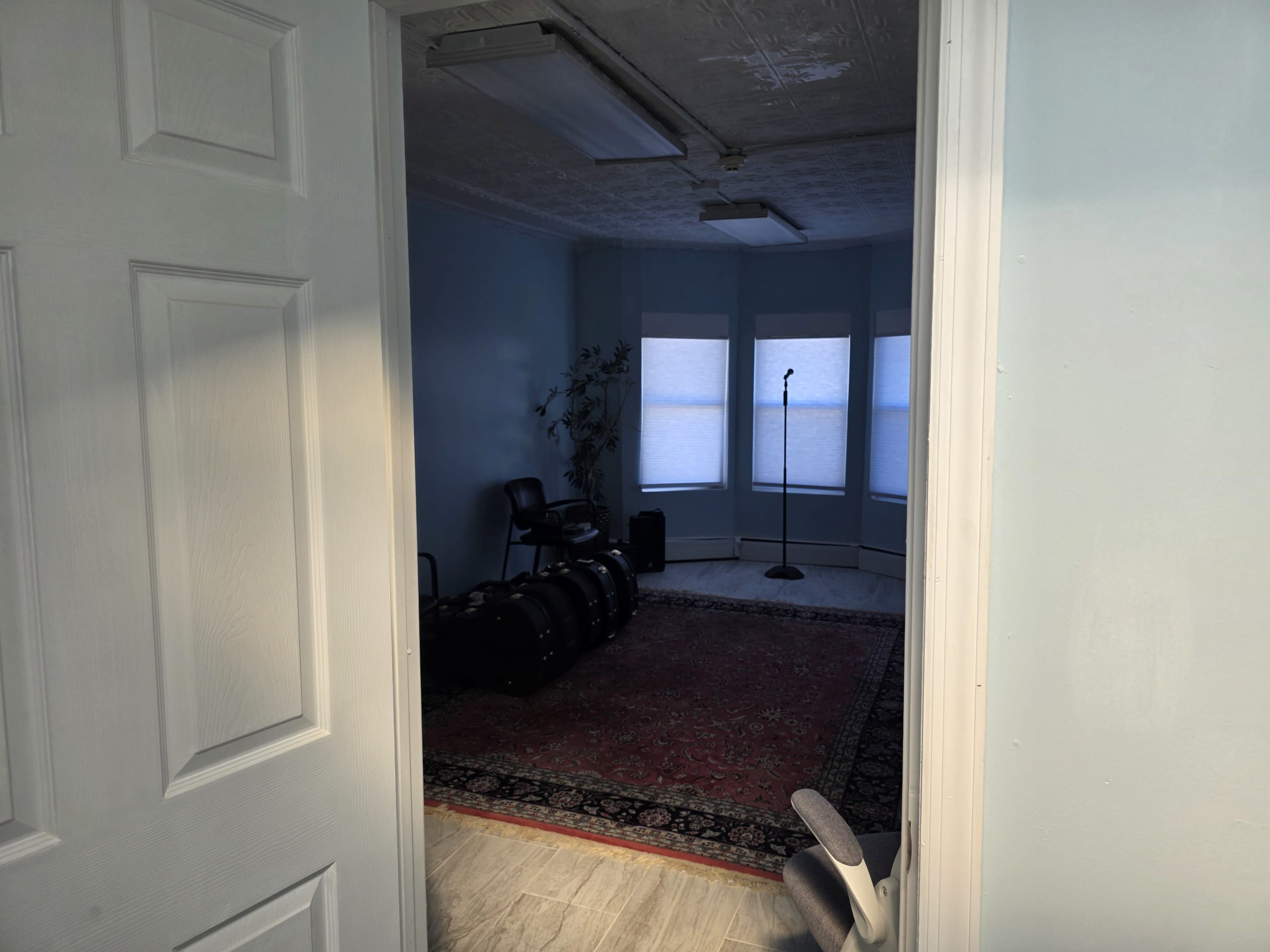 The image shows a room with light blue walls, a microphone stand, chairs, and several black cases on a red carpet.