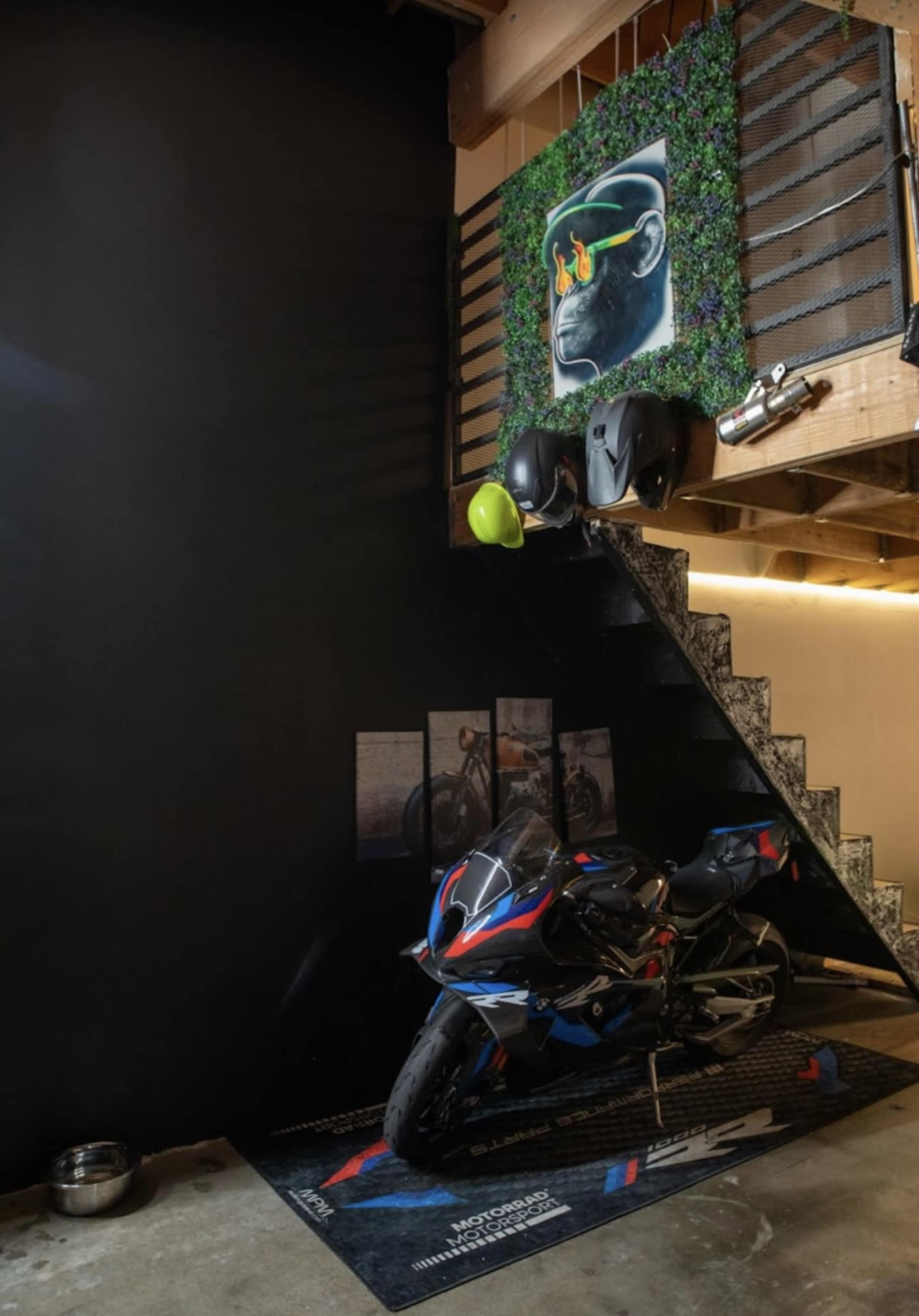 A motorcycle is parked at the base of a staircase, accompanied by two helmets, and there is a large artwork on the wall above decorated with greenery.