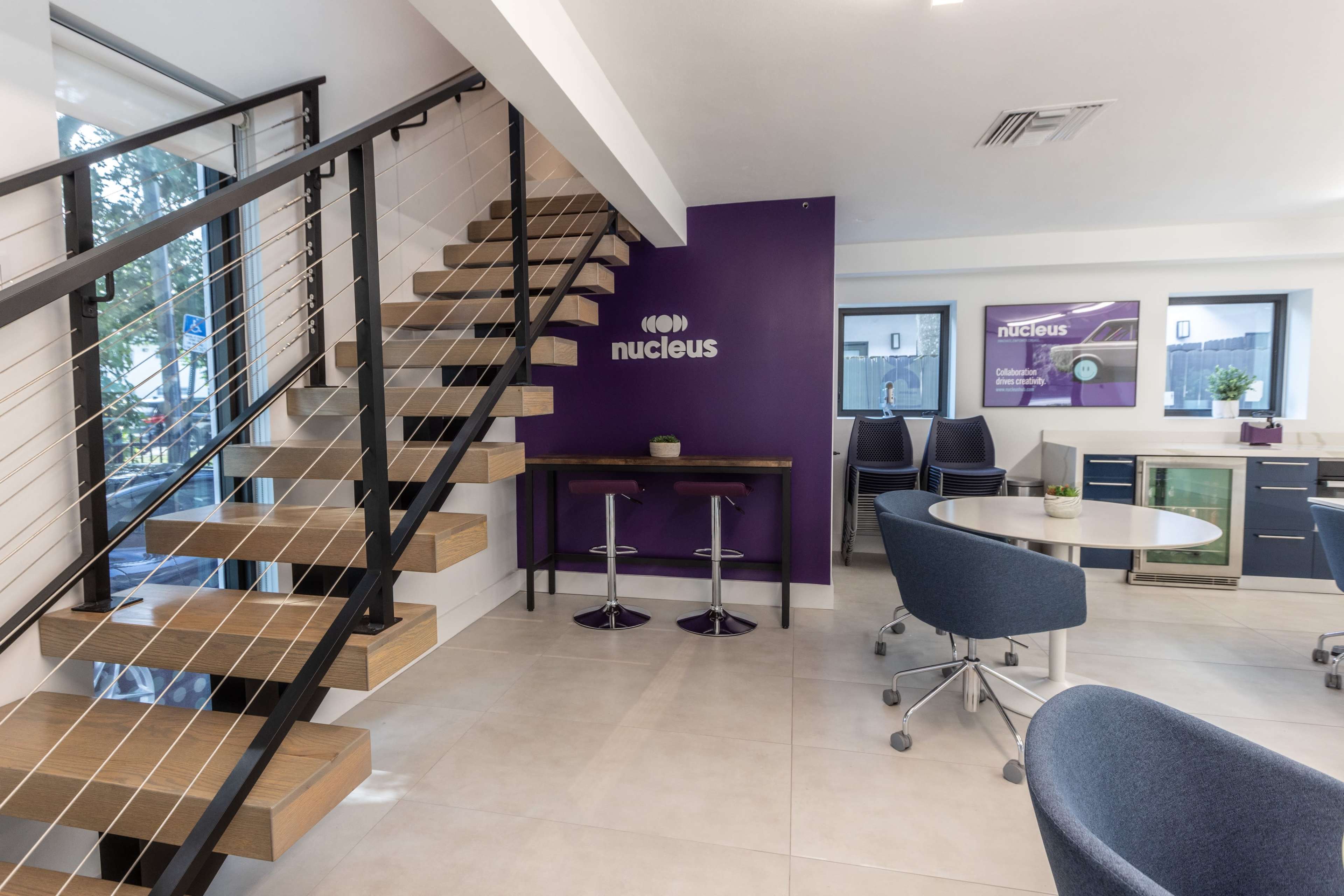 A modern interior space featuring a staircase, a bar with stools, and a seating area, with purple accent walls and contemporary furniture.