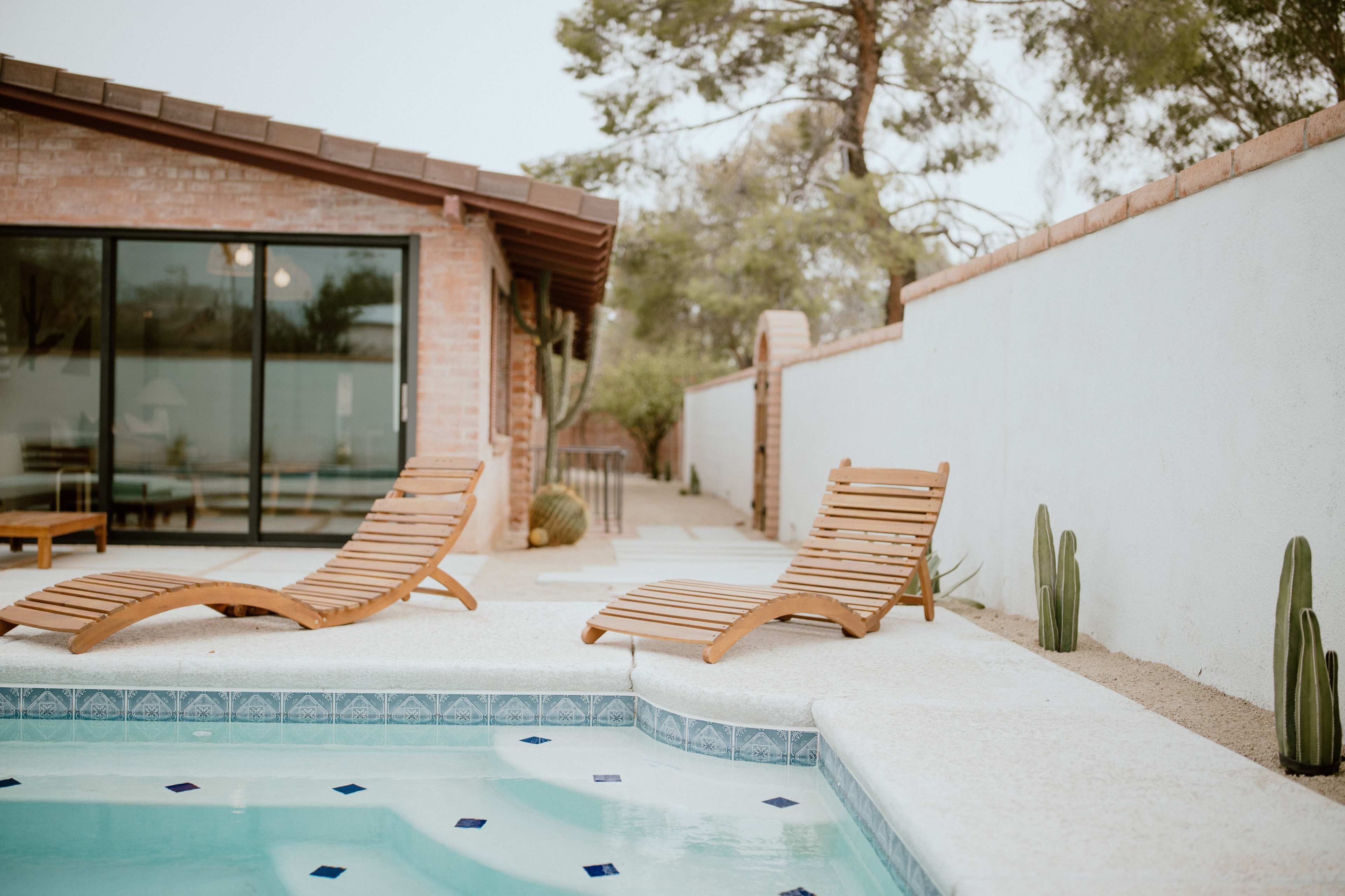 Two wooden lounge chairs rest by a swimming pool in a landscaped backyard, with a wall and cacti in the background.