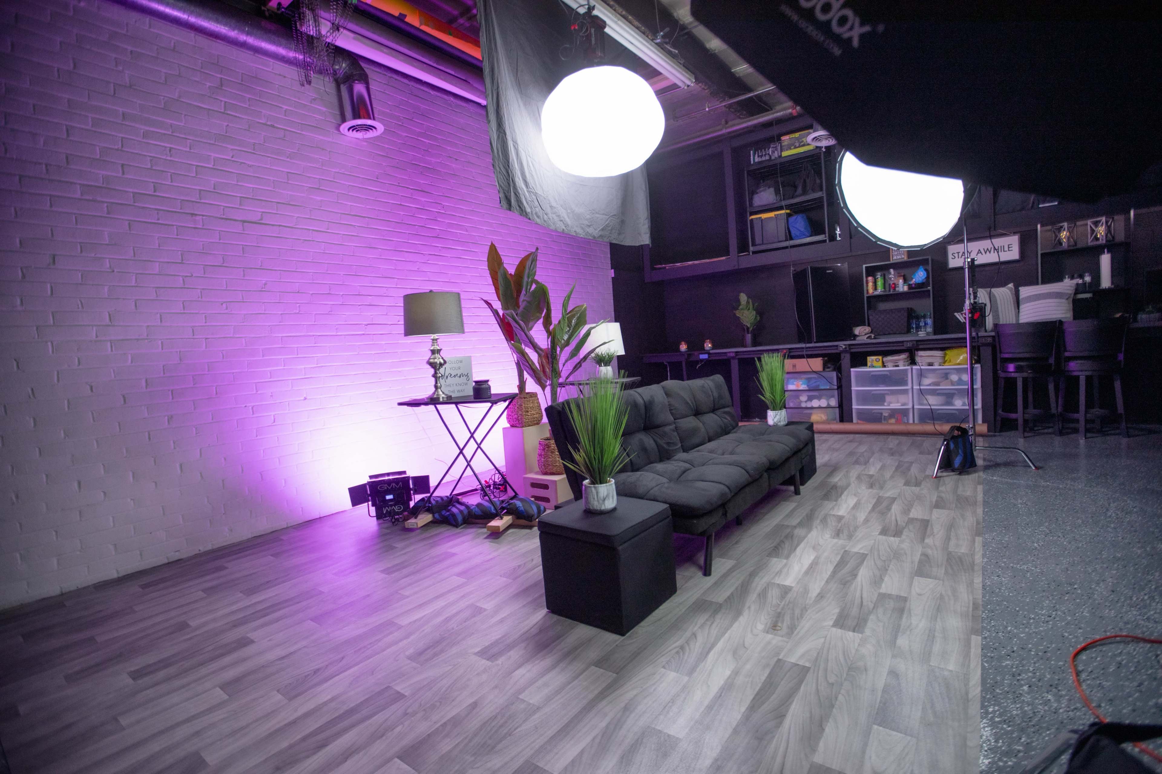 A modern studio space featuring a black couch, a lamp, a small table, and decorative plants, with soft purple lighting against a textured white wall.