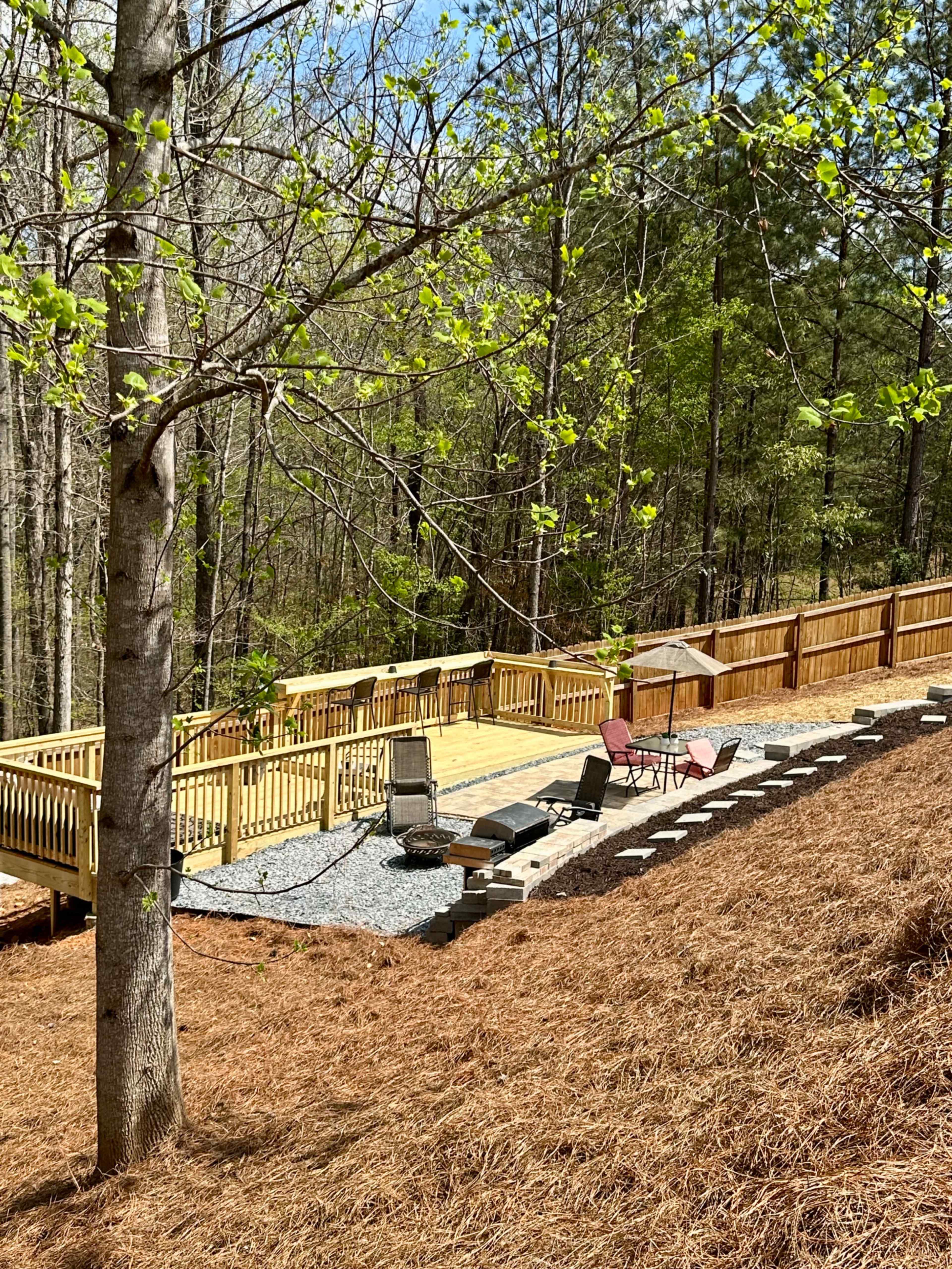 A wooden deck with lounge chairs and a small table is surrounded by trees and a sloped yard covered with pine needles.