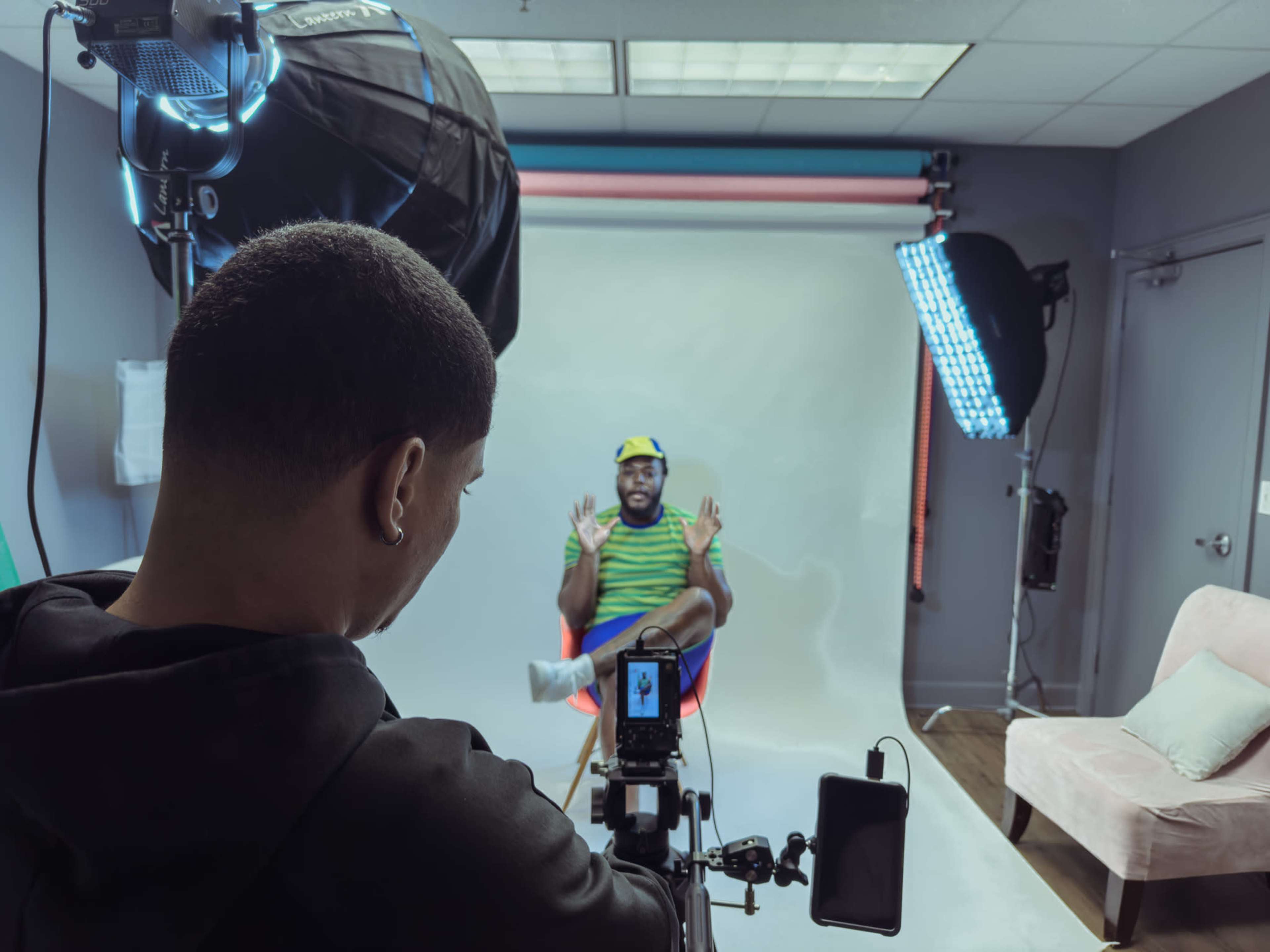A person is filming an interview in a studio setting with a subject sitting in a chair against a backdrop.