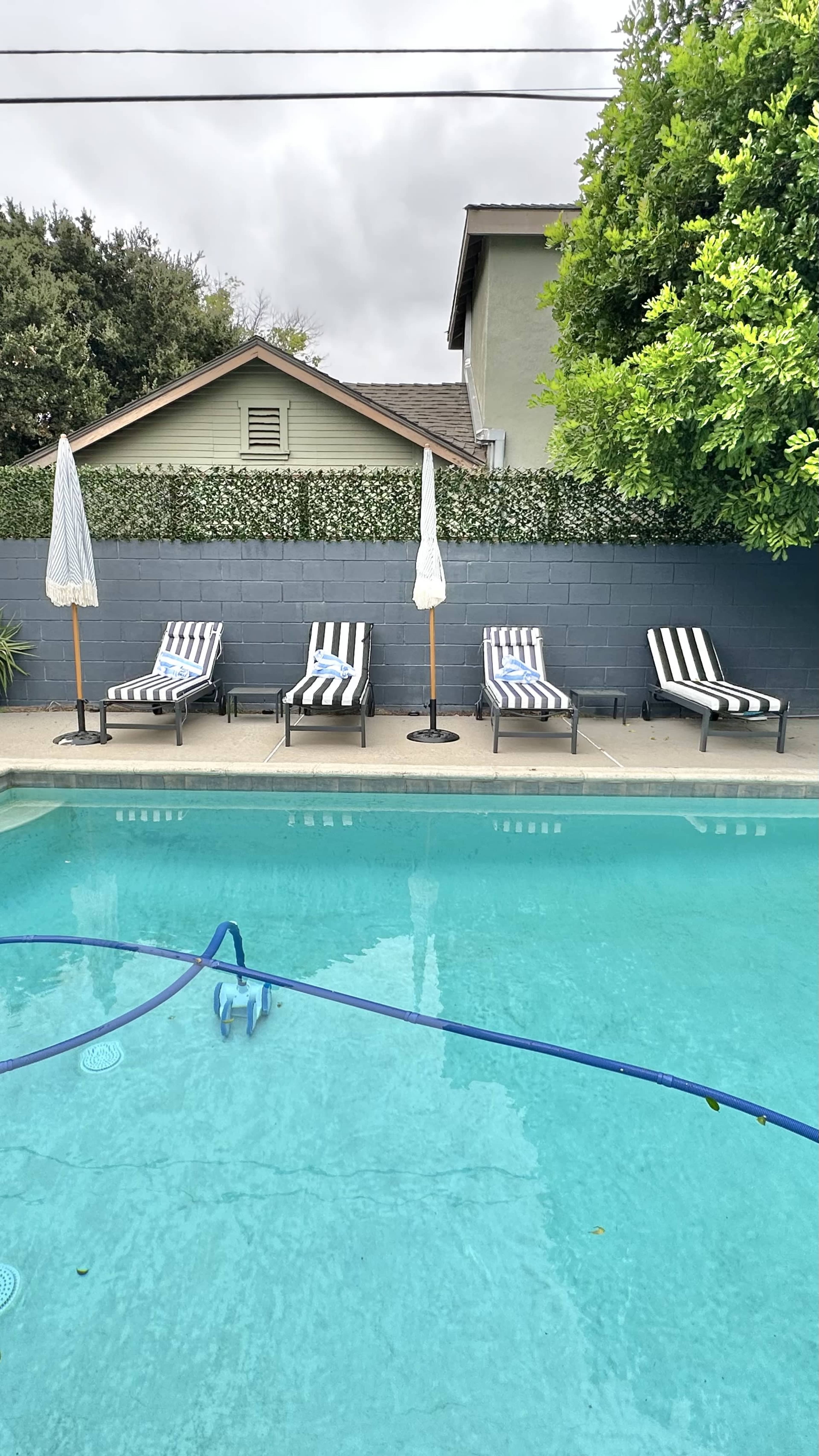 Lounge chairs with striped cushions are arranged alongside a swimming pool, separated by a blue wall and two umbrellas.