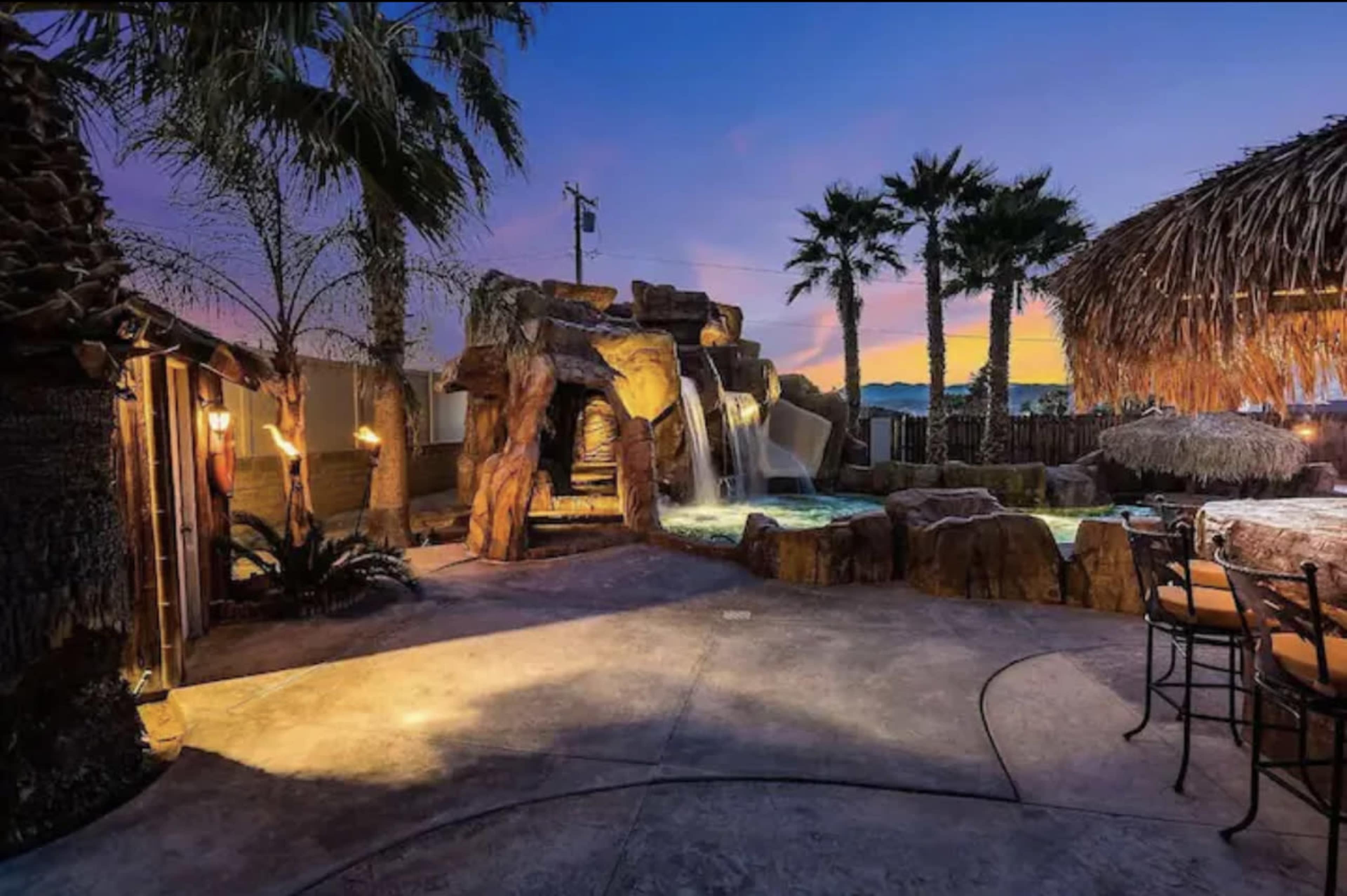 A backyard oasis featuring a rock waterfall that flows into a pool, surrounded by palm trees and a tiki hut bar, illuminated by twilight.