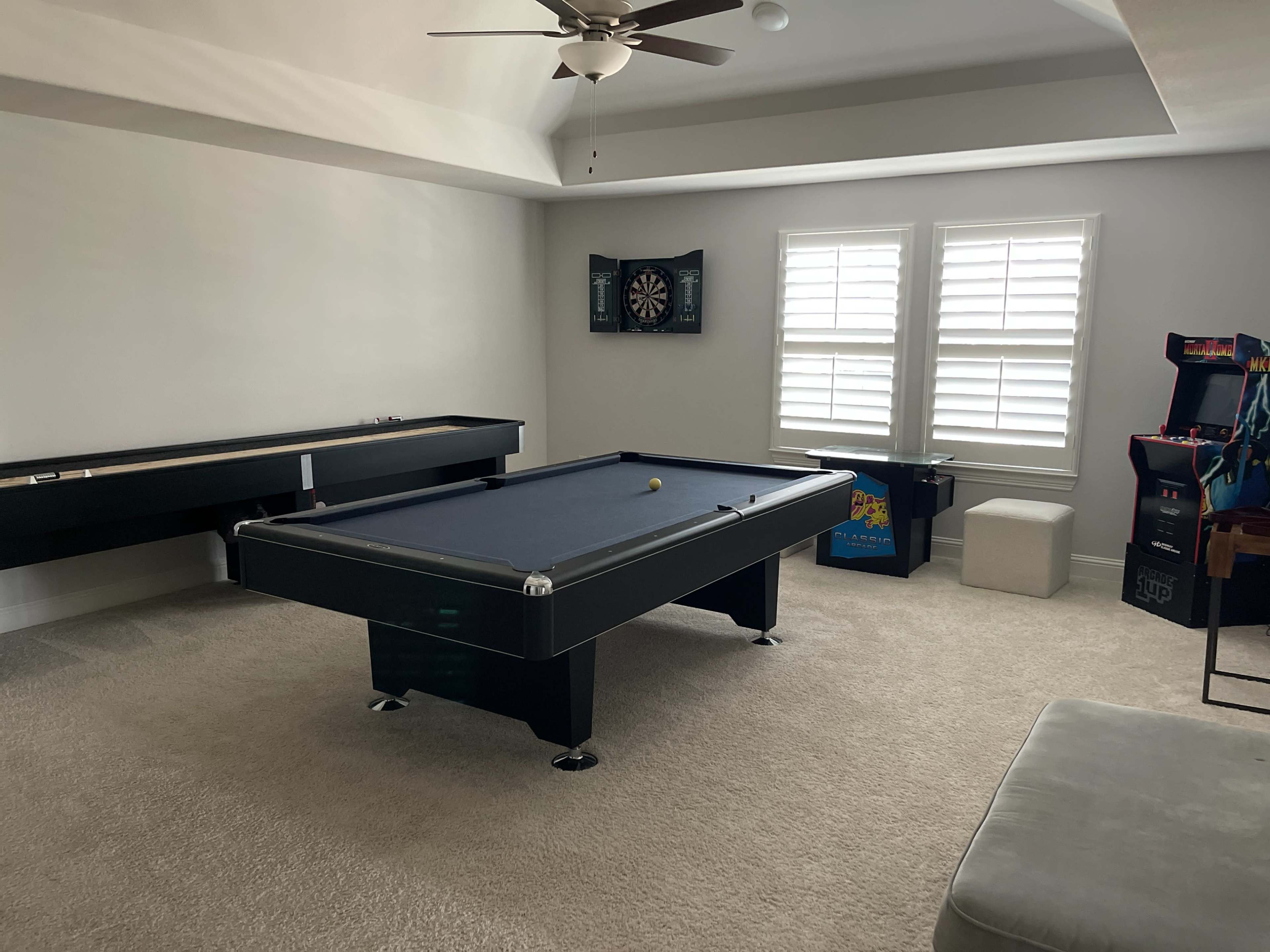 A game room with a pool table, dartboard, arcade machine, and a cozy seating area.