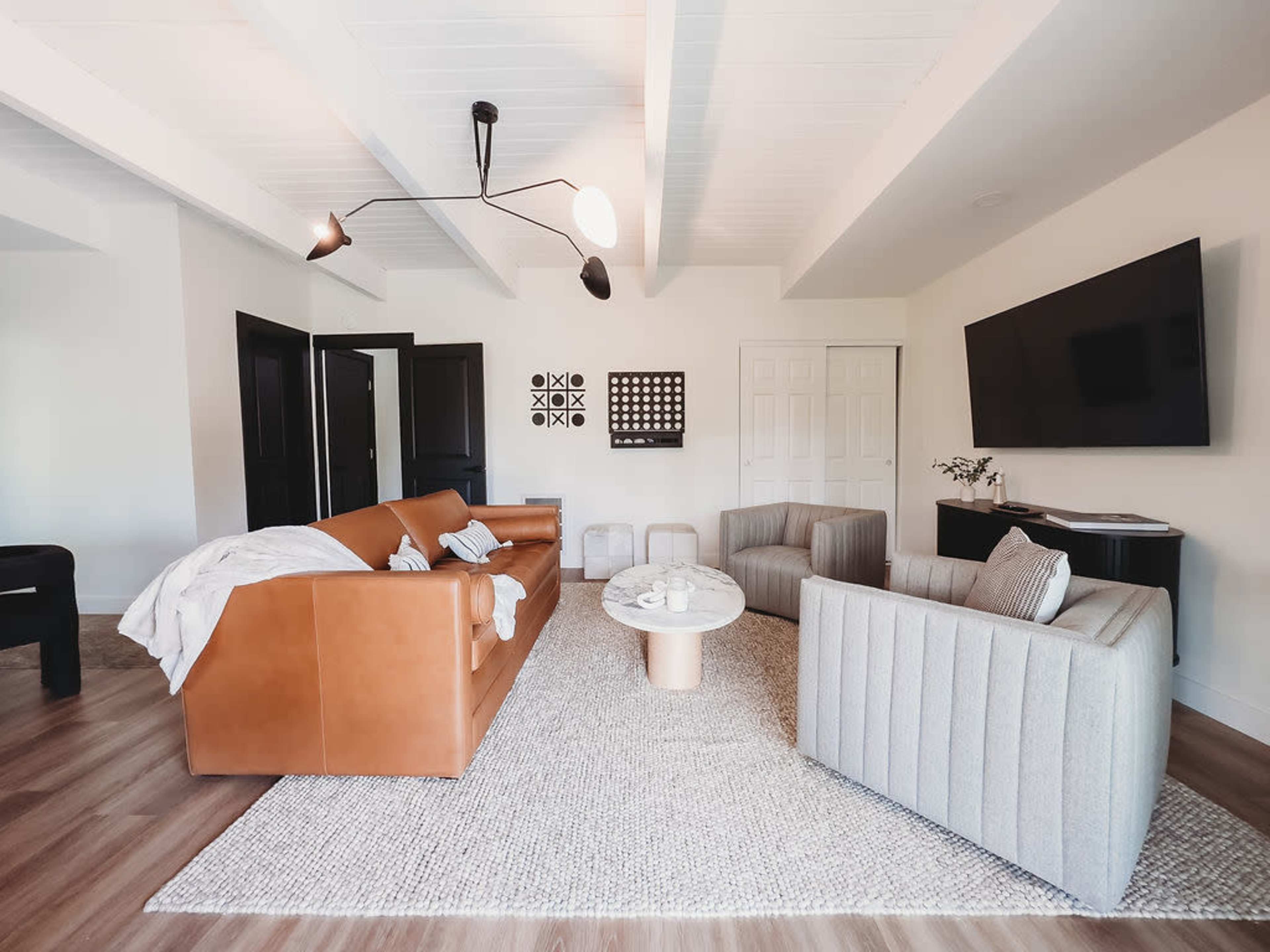 The image shows a modern living room featuring a brown leather sofa, two light gray armchairs, a round coffee table, and a large flat-screen TV mounted on the wall.