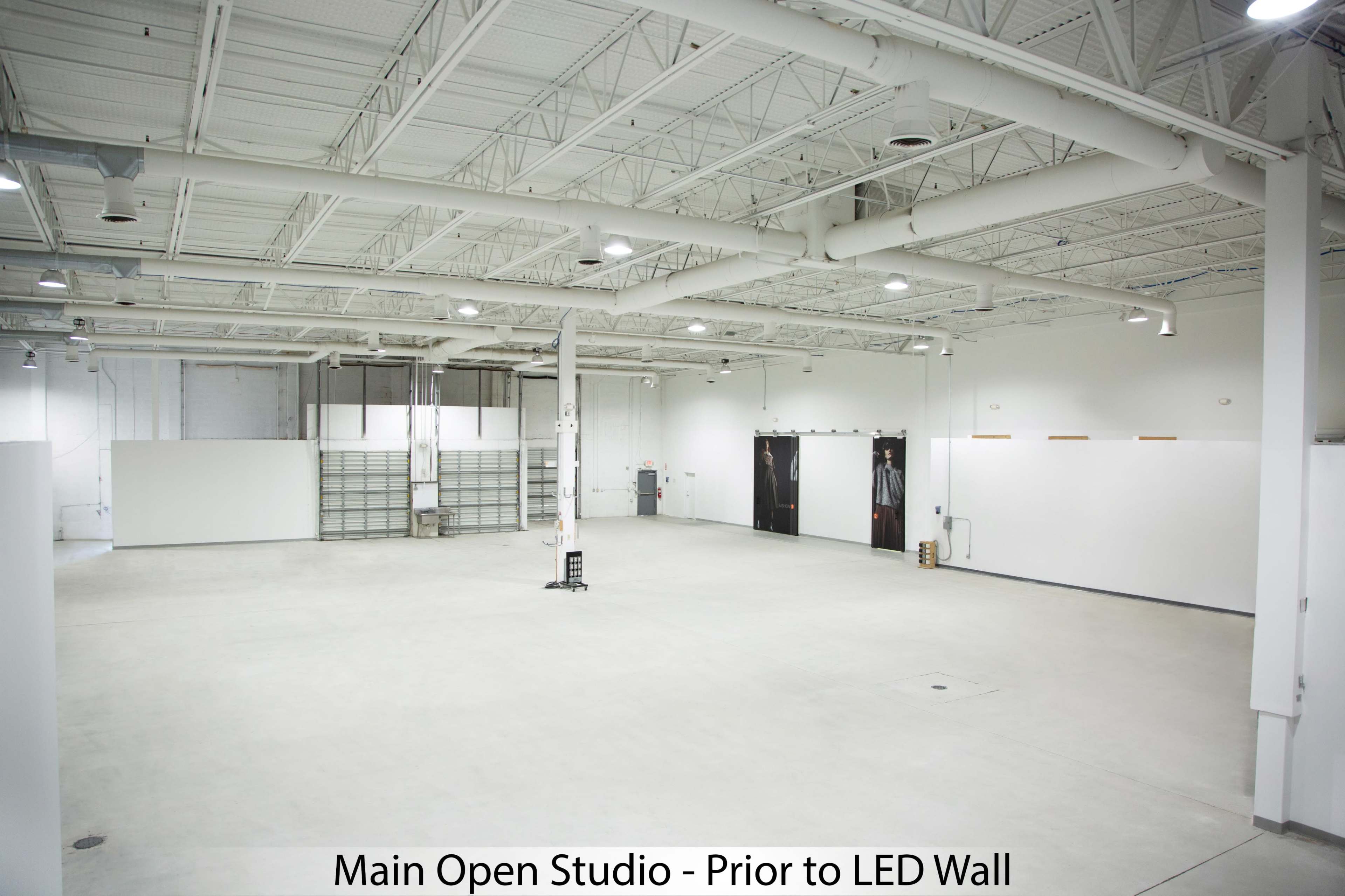 The image shows a large, empty open studio space with concrete flooring and white walls, featuring overhead lighting and a few utility elements.