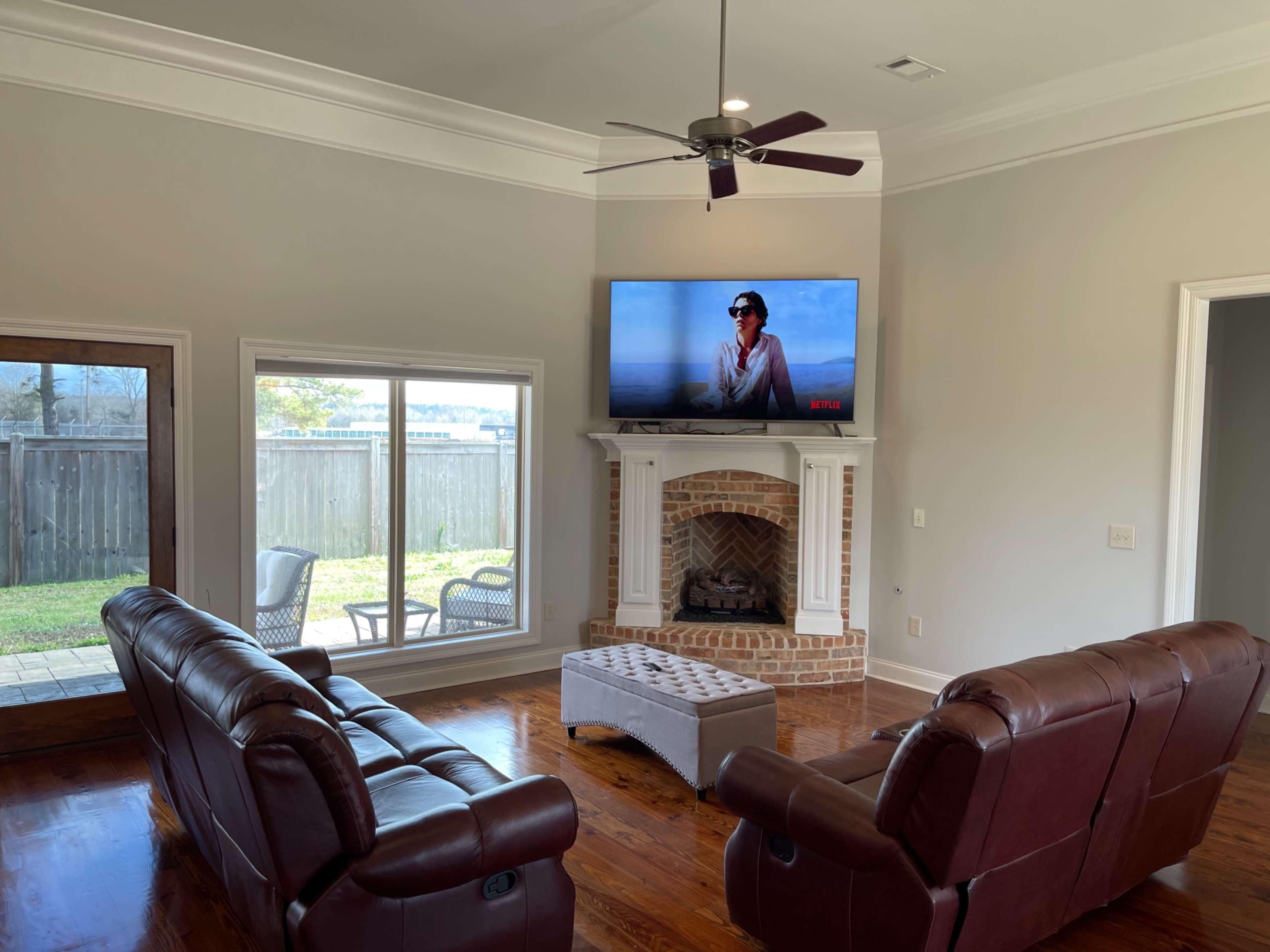 A living room features a TV above a brick fireplace, with two leather reclining couches facing it and a view of a backyard through a large window.
