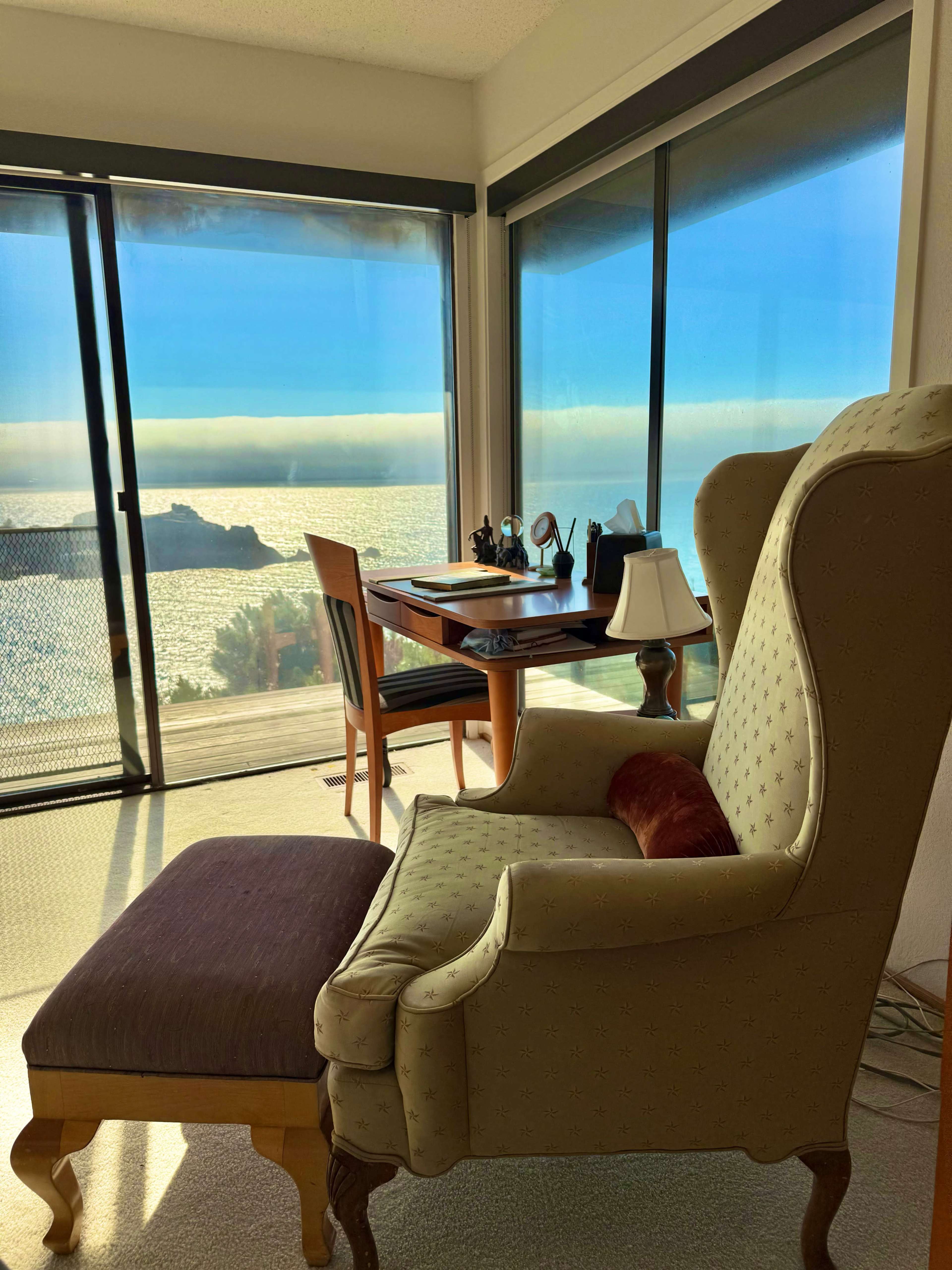 A cozy chair and ottoman are positioned near a large window with a view of the ocean and a desk in the background.