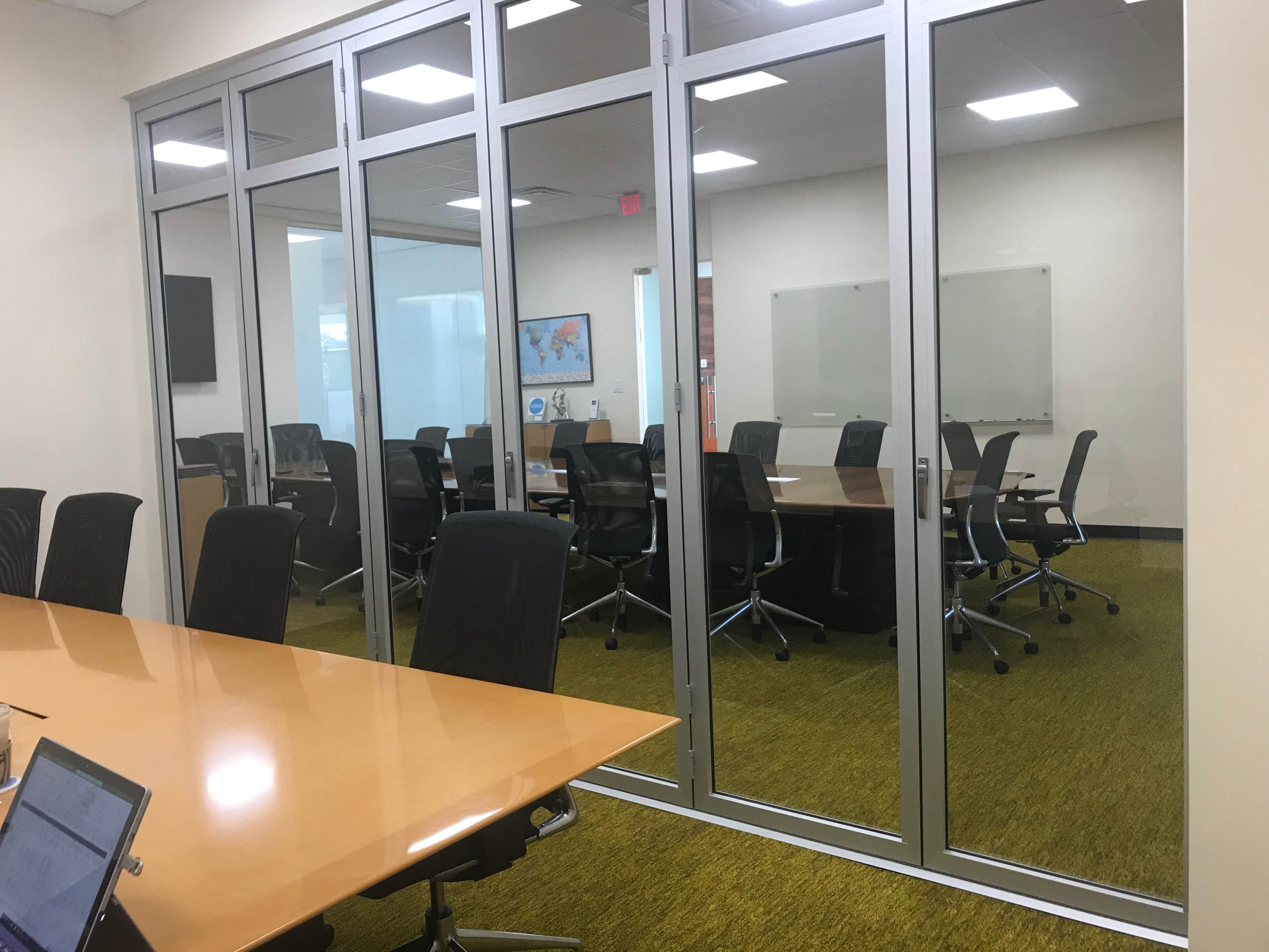 A glass-walled conference room features a long wooden table surrounded by swivel chairs and is visible through transparent walls.