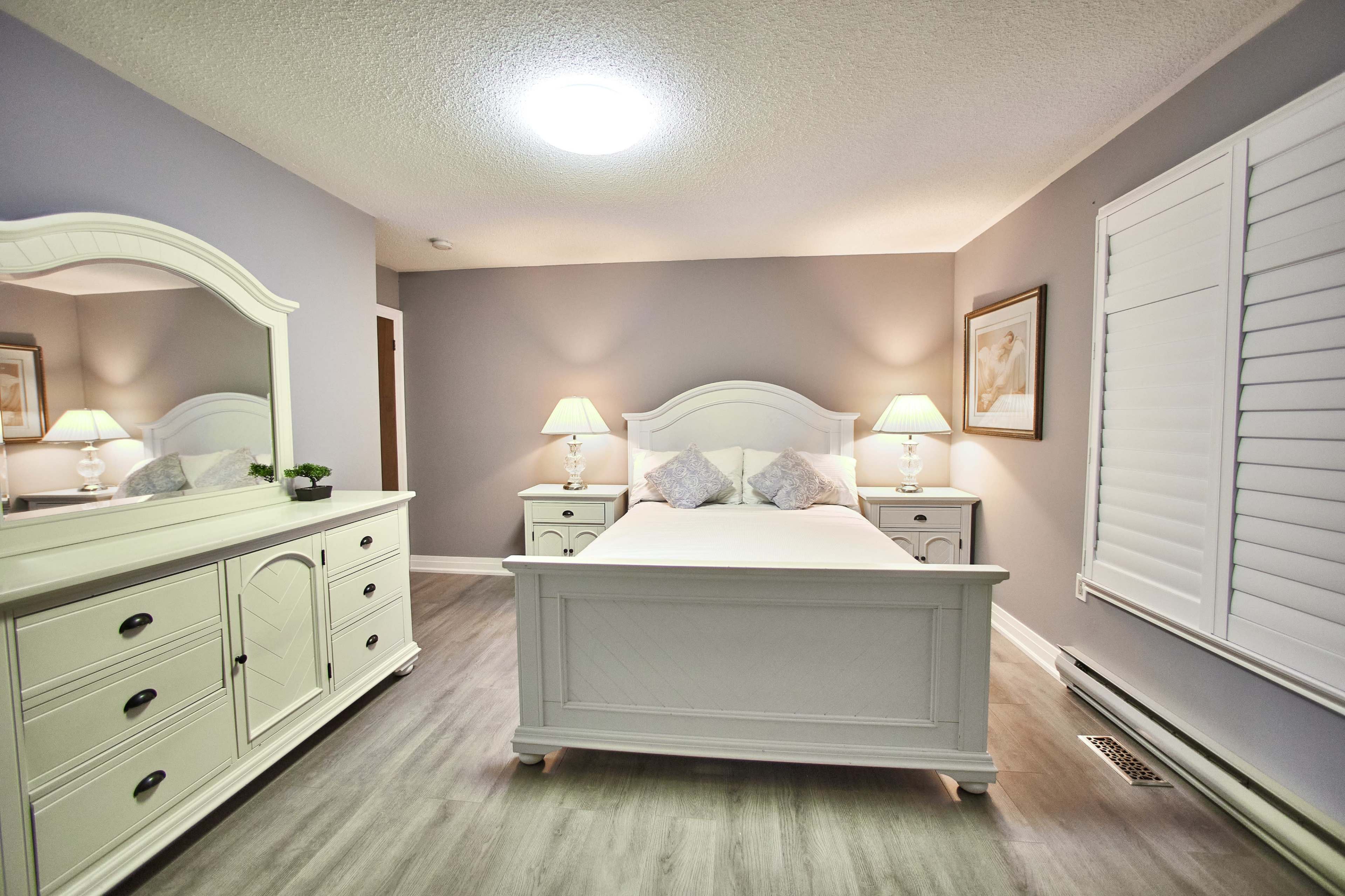 A well-lit bedroom features a white bed frame, matching nightstands, and a dresser against pale gray walls.