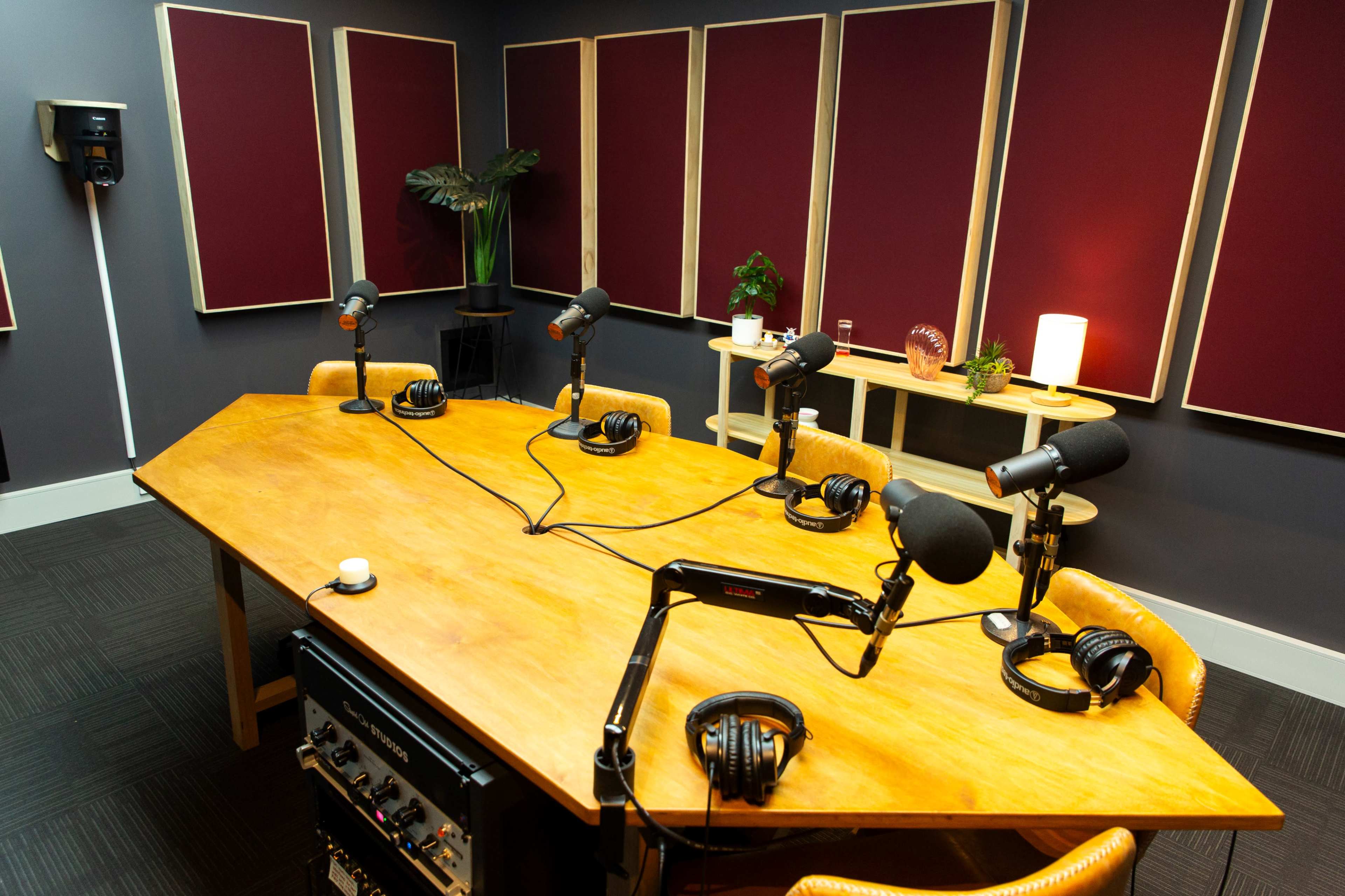 A recording studio features a large wooden table with six microphones and headphones arranged around it, alongside soundproofing panels on the walls.