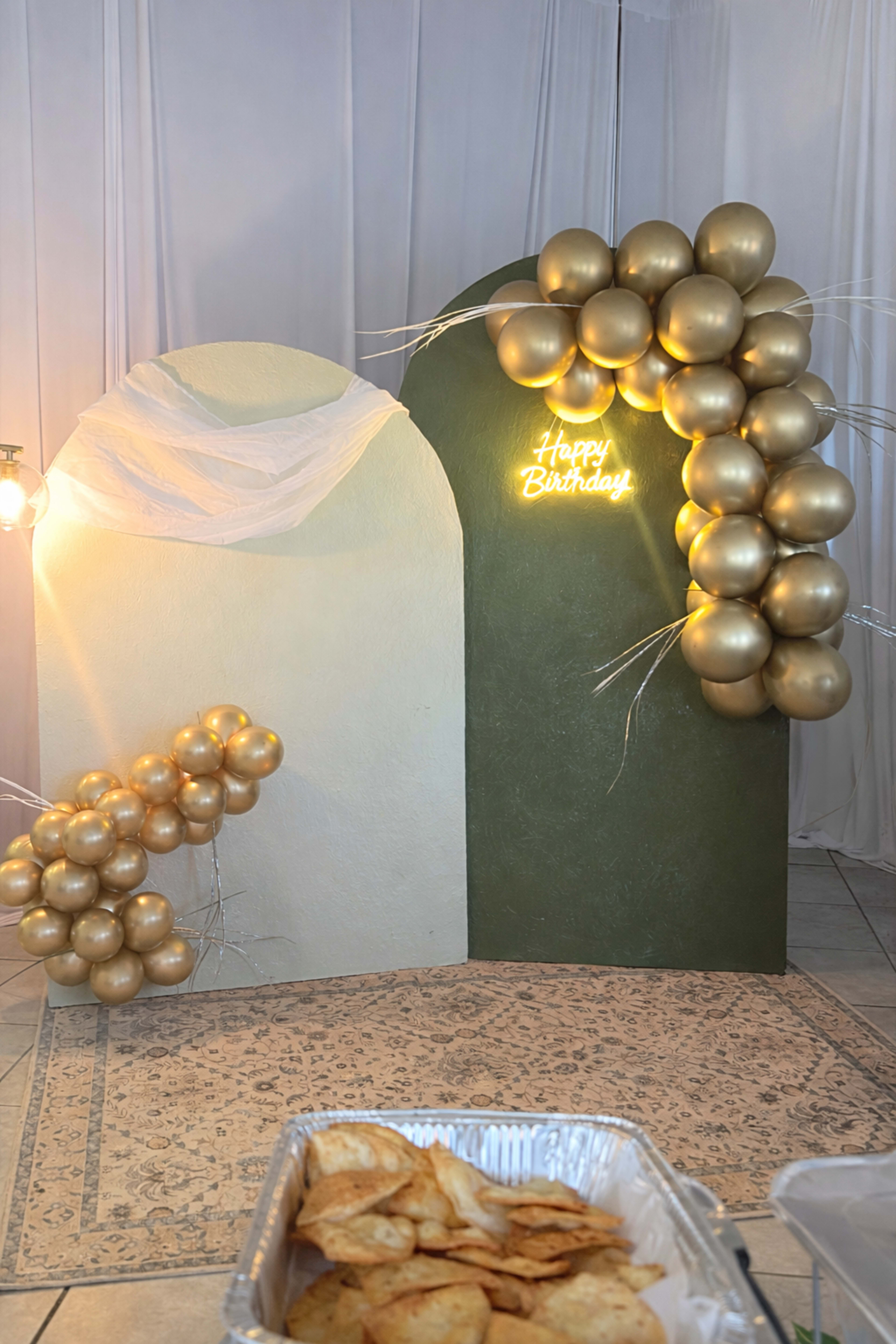 The image shows a decorative birthday backdrop featuring gold balloons arranged in an arch beside a "Happy Birthday" neon sign, with a tray of snacks in the foreground.