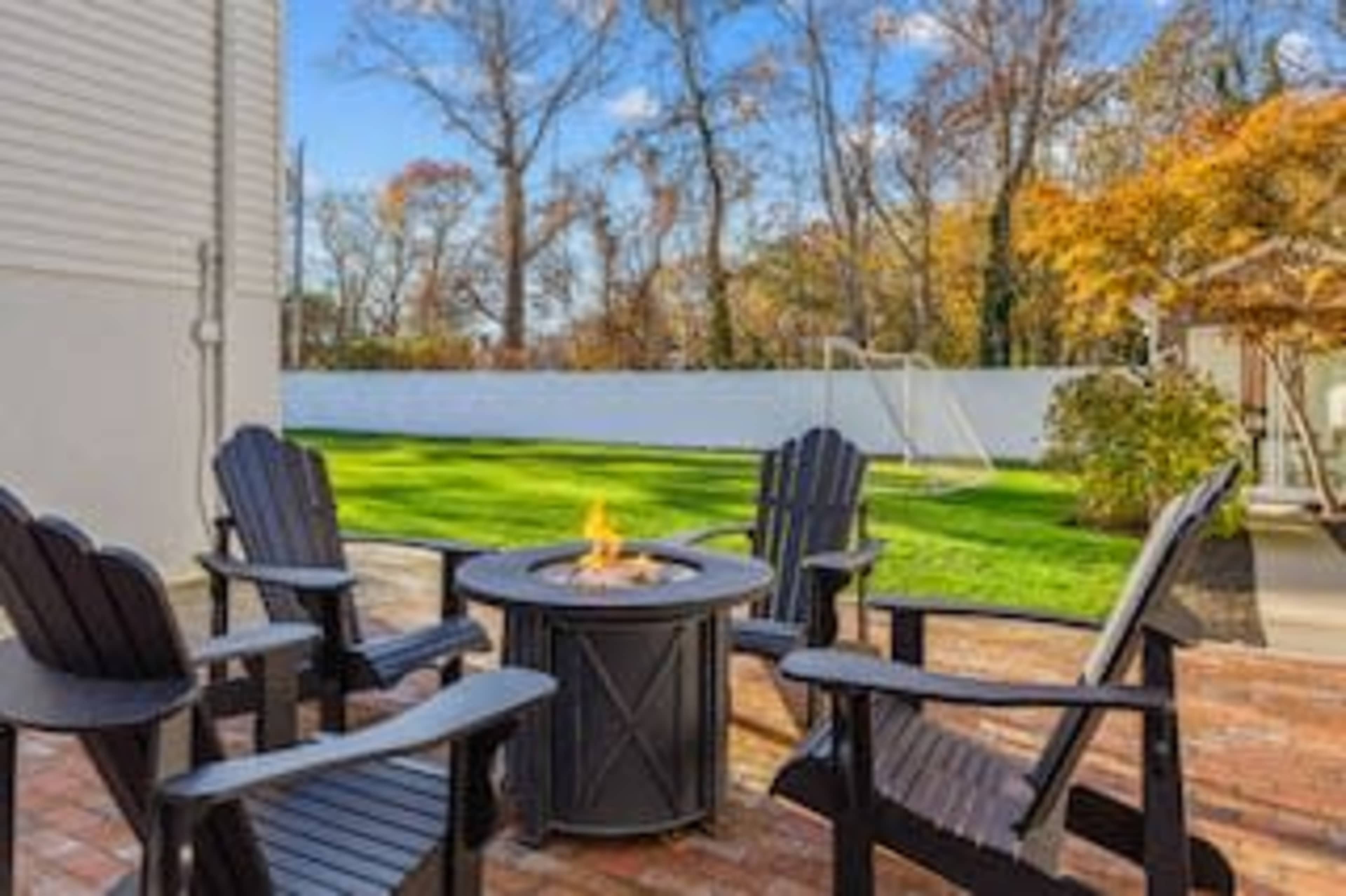 A circular fire pit surrounded by four wooden chairs stands on a brick patio, overlooking a grassy yard with trees in the background.