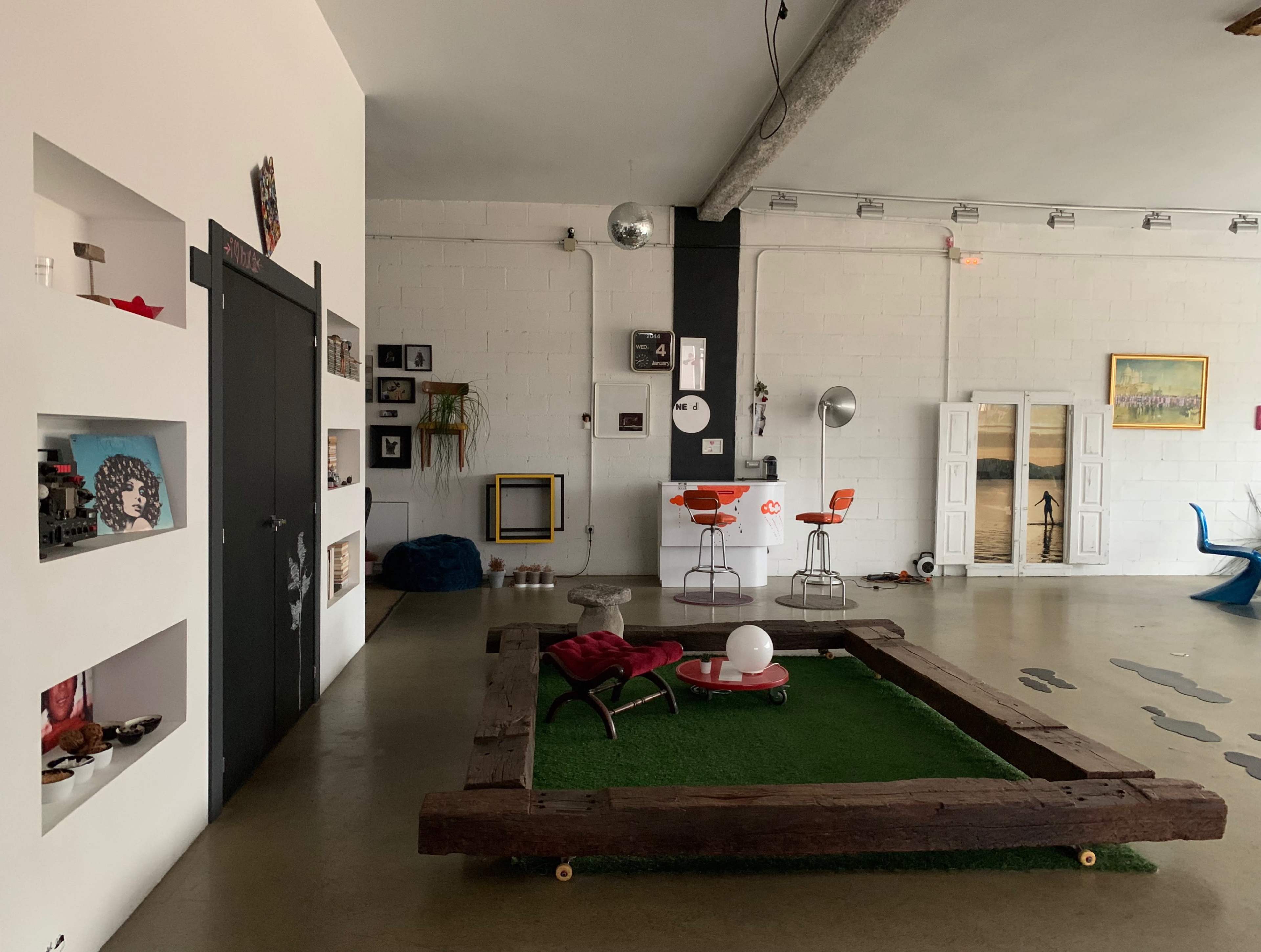 A spacious, modern loft interior featuring a wooden coffee table on grass, eclectic decor on the walls, and a small kitchen area with orange bar stools.