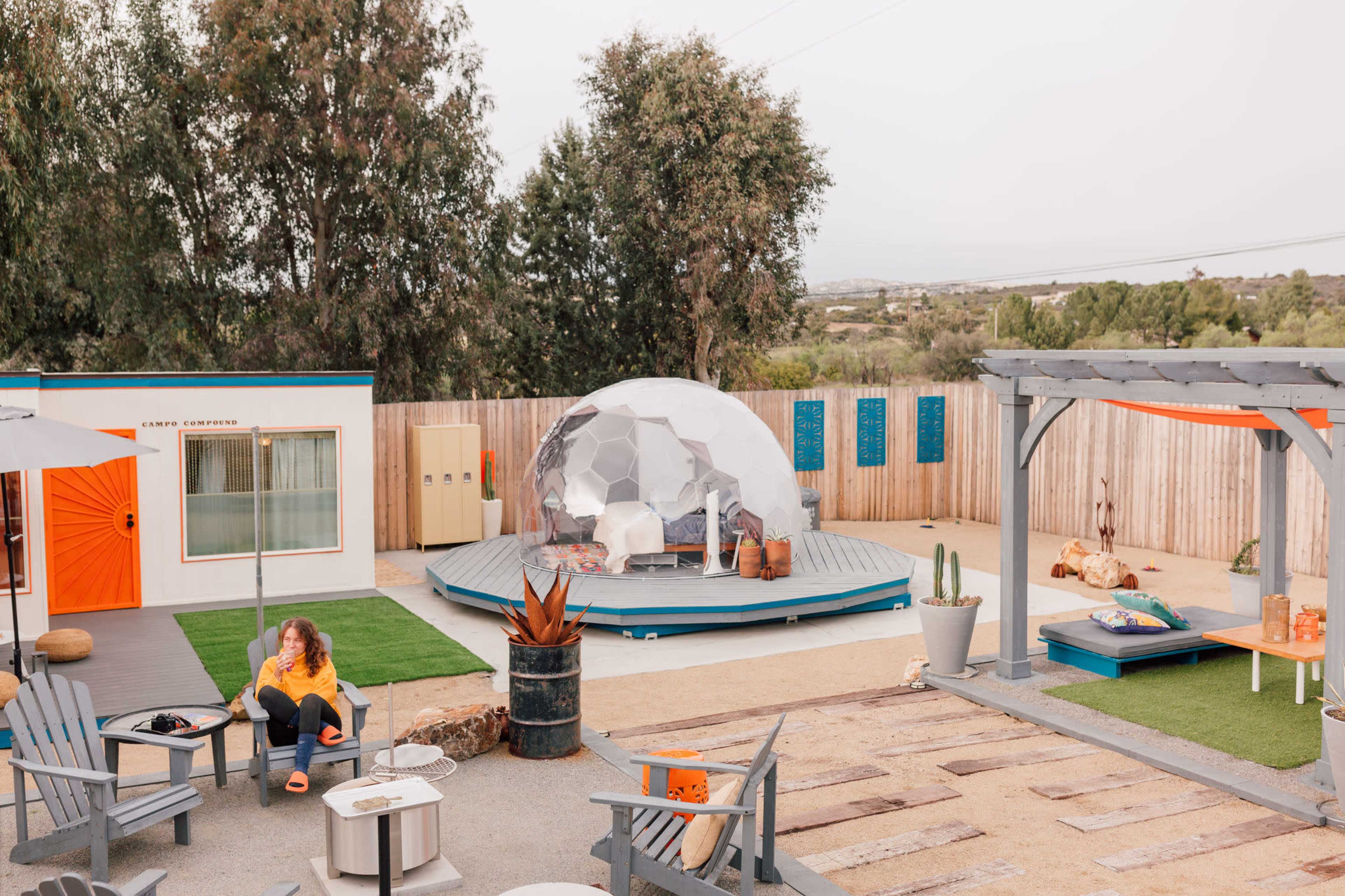 A modern outdoor space featuring a geodesic dome, wooden seating areas, and a gravel pathway, surrounded by greenery and a wooden fence.