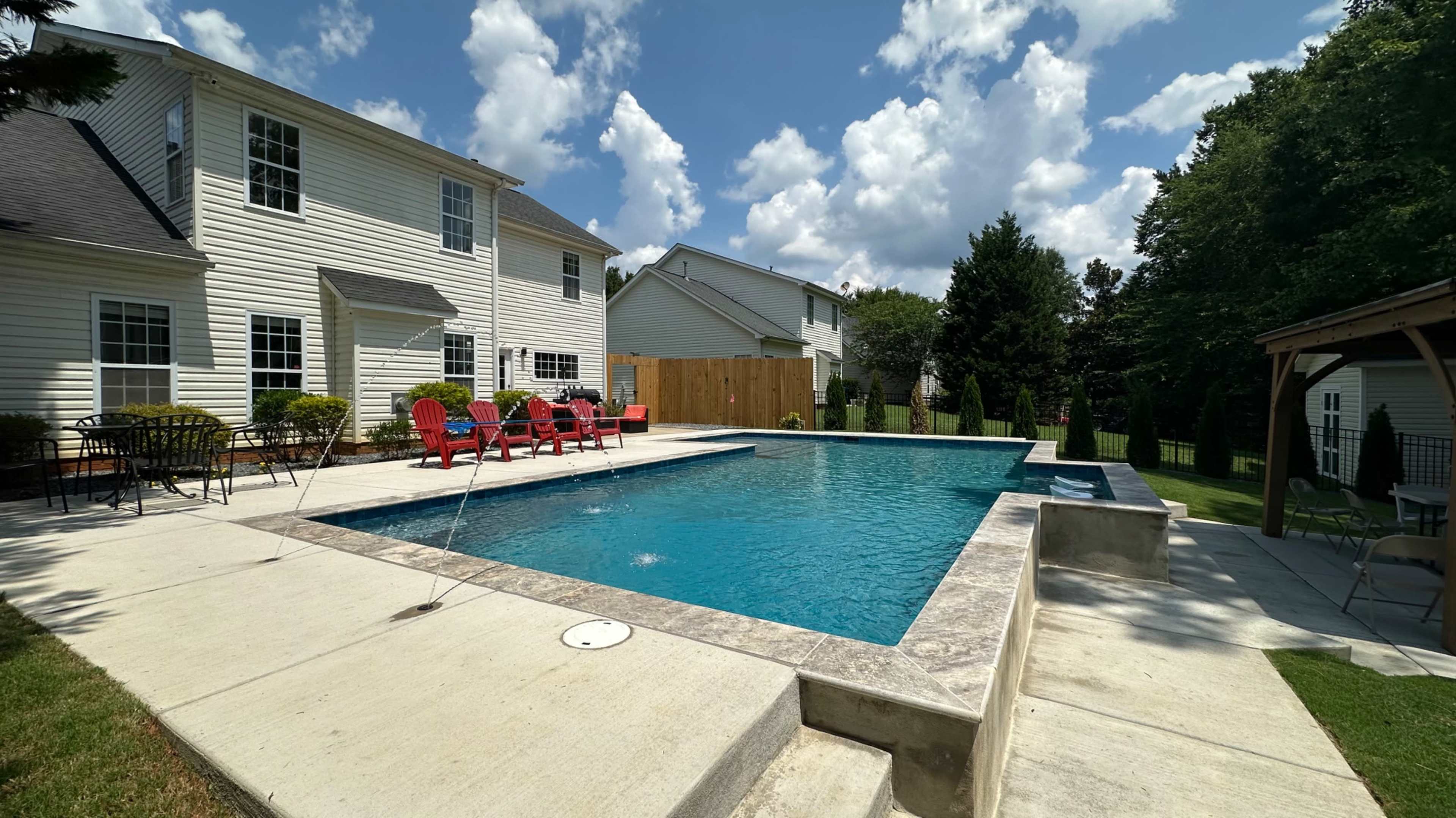 A rectangular swimming pool is surrounded by a concrete patio and lounge chairs in a backyard, with a fence and trees in the background under a partly cloudy sky.