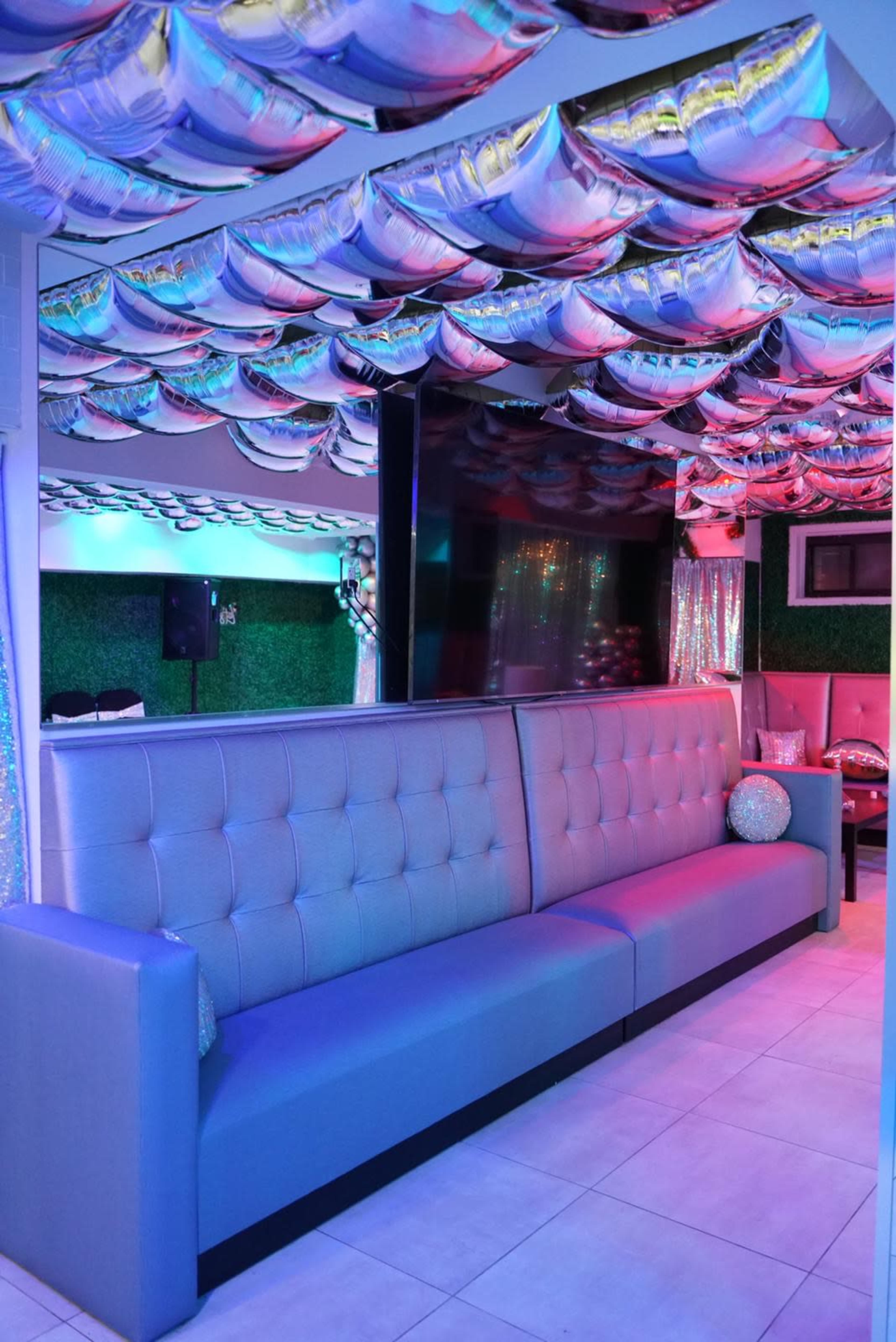 A lounge area with a gray upholstered bench, reflecting colorful lights from metallic balloon-like decorations on the ceiling.