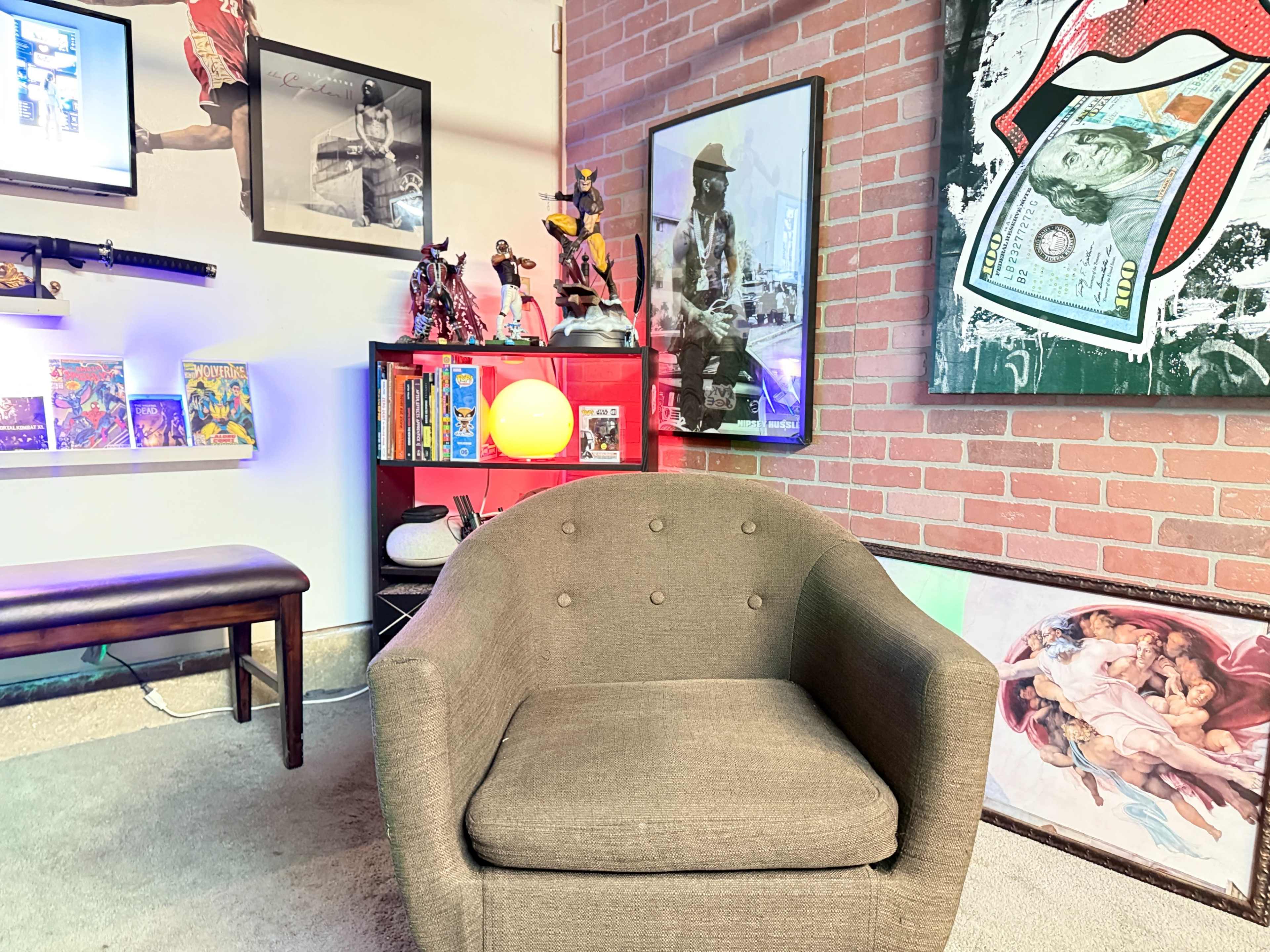 The image shows a cozy lounge area featuring a brown upholstered chair, a brick wall, various framed artworks, and shelves displaying collectibles and books.