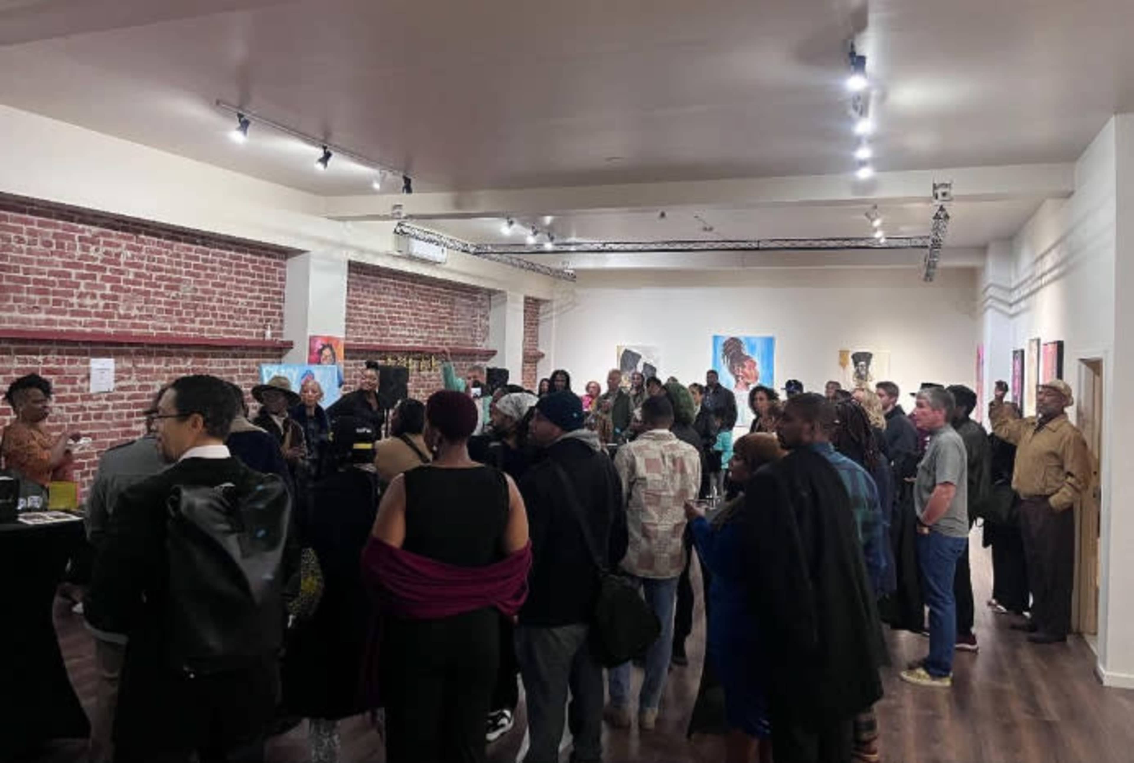 A large group of people gathers in a gallery space filled with artwork and exposed brick walls.
